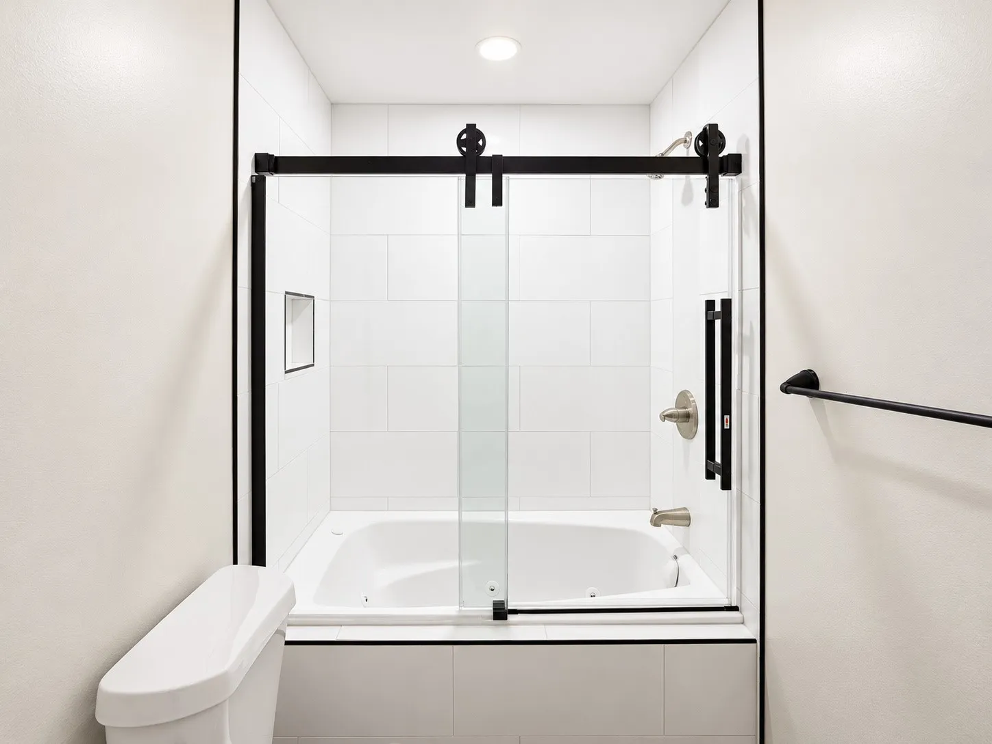 White tile surround bathtub with black framed sliding glass door, brushed nickel fixtures, and recessed shelf