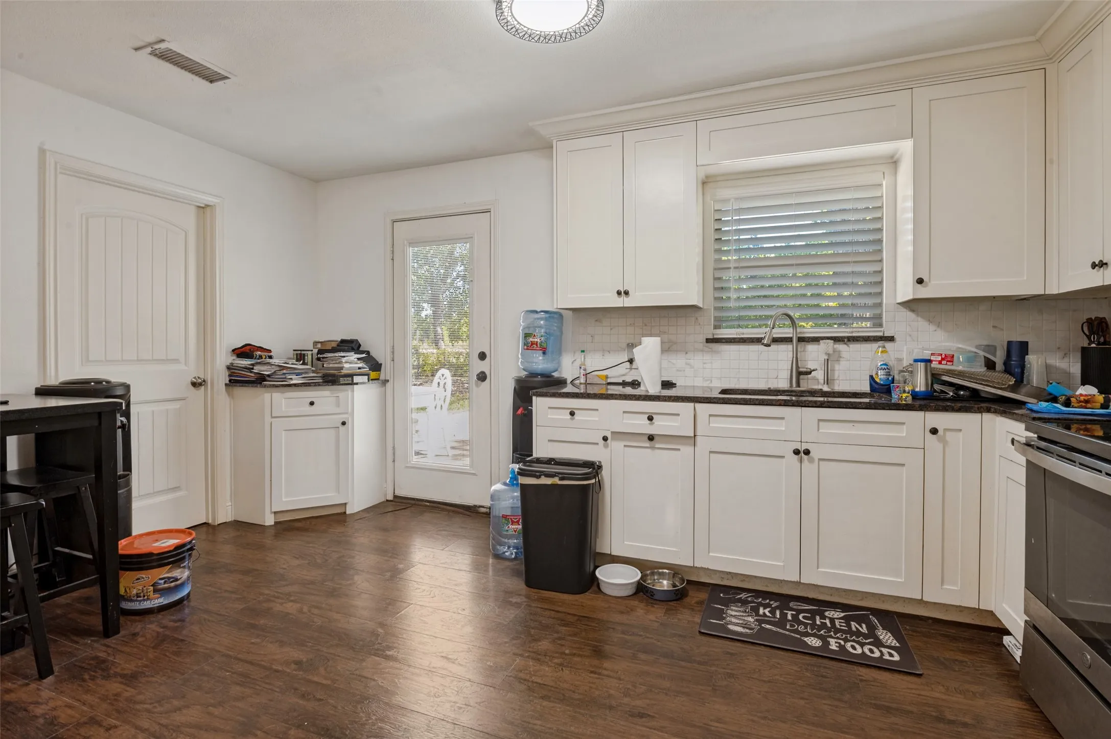 White shaker-style cabinetry with dark hardware, dark countertops, a tile backsplash, stainless steel sink with a gooseneck faucet, and wood-finish flooring