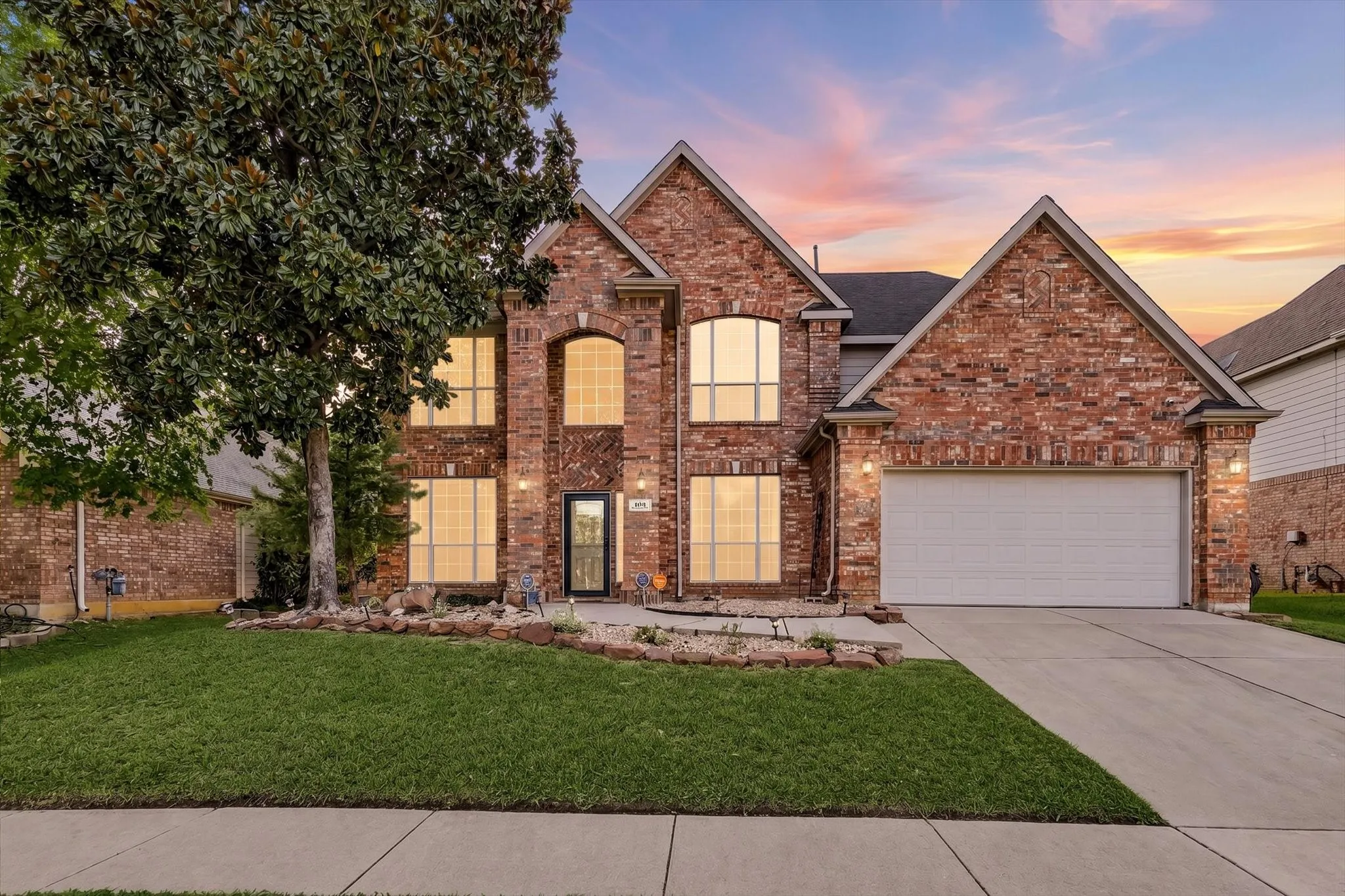 Brick exterior with multiple gables, arched entry, mature front yard tree, concrete driveway, and attached garage
