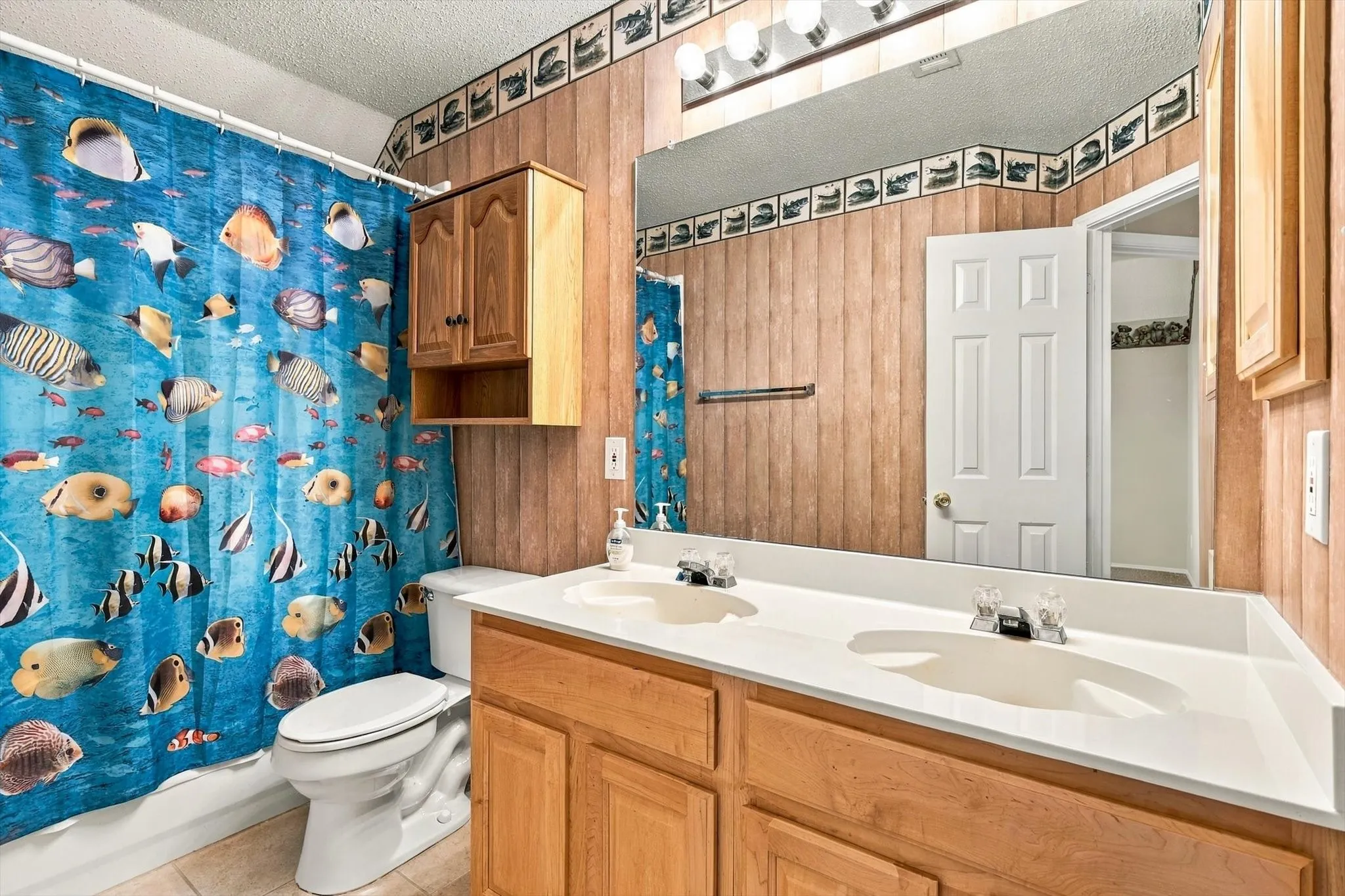 Dual sink vanity with light wood-finish cabinetry, a large wall-mounted mirror, and overhead vanity lighting