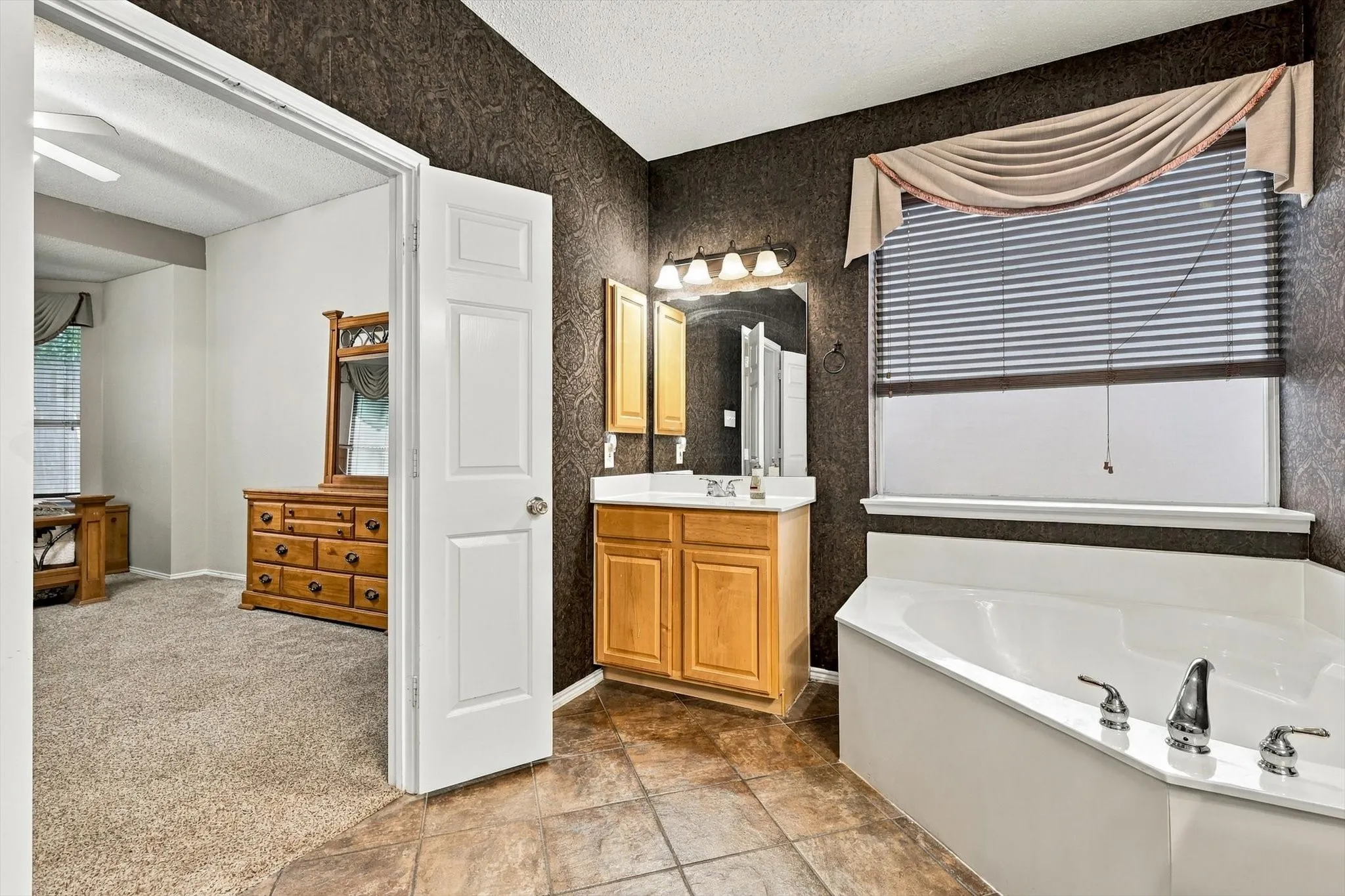 Bathroom featuring a built-in corner soaking tub, a single vanity with wood-finish cabinetry, and patterned wall covering