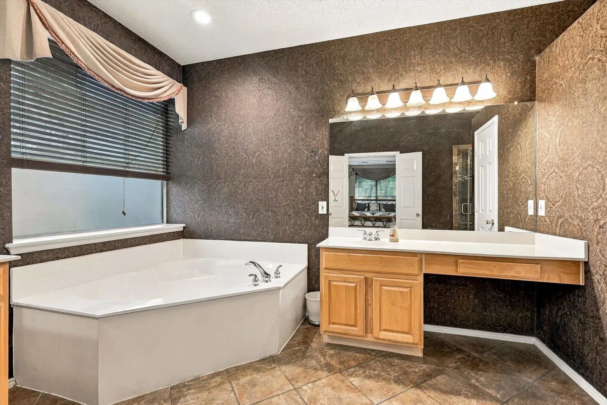 Corner soaking tub with deck-mounted fixtures, wood-finish vanity with countertop, large wall-mounted mirror, and tile flooring