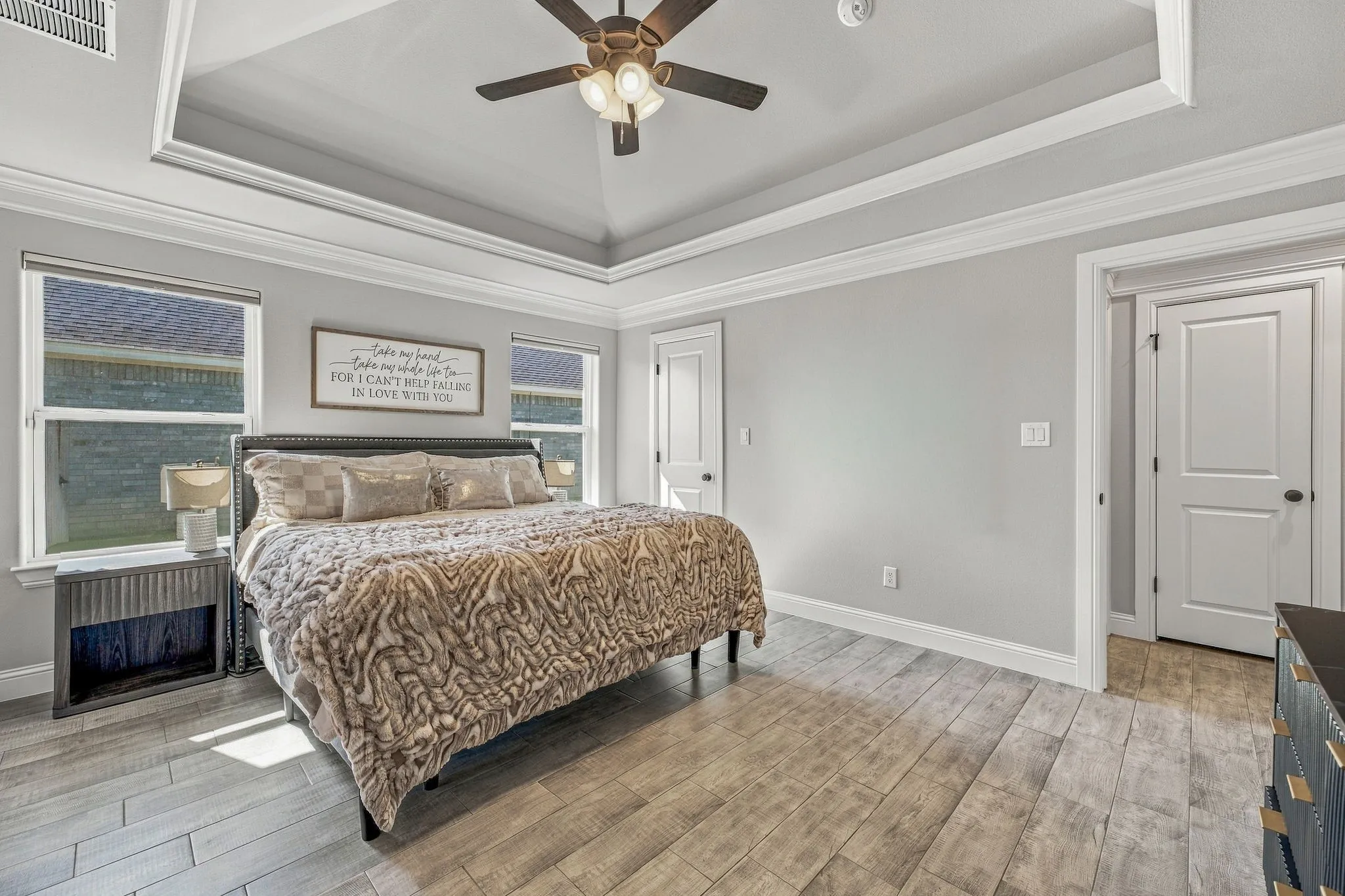 Tray ceiling with crown molding, ceiling fan, two windows with blinds, wood-finish flooring, and white interior doors