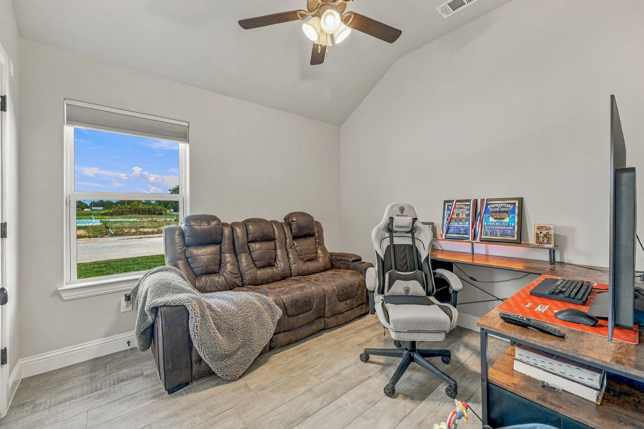 Vaulted ceiling with a ceiling fan, light gray wall paint, wood-finish flooring, and a window with roller shade