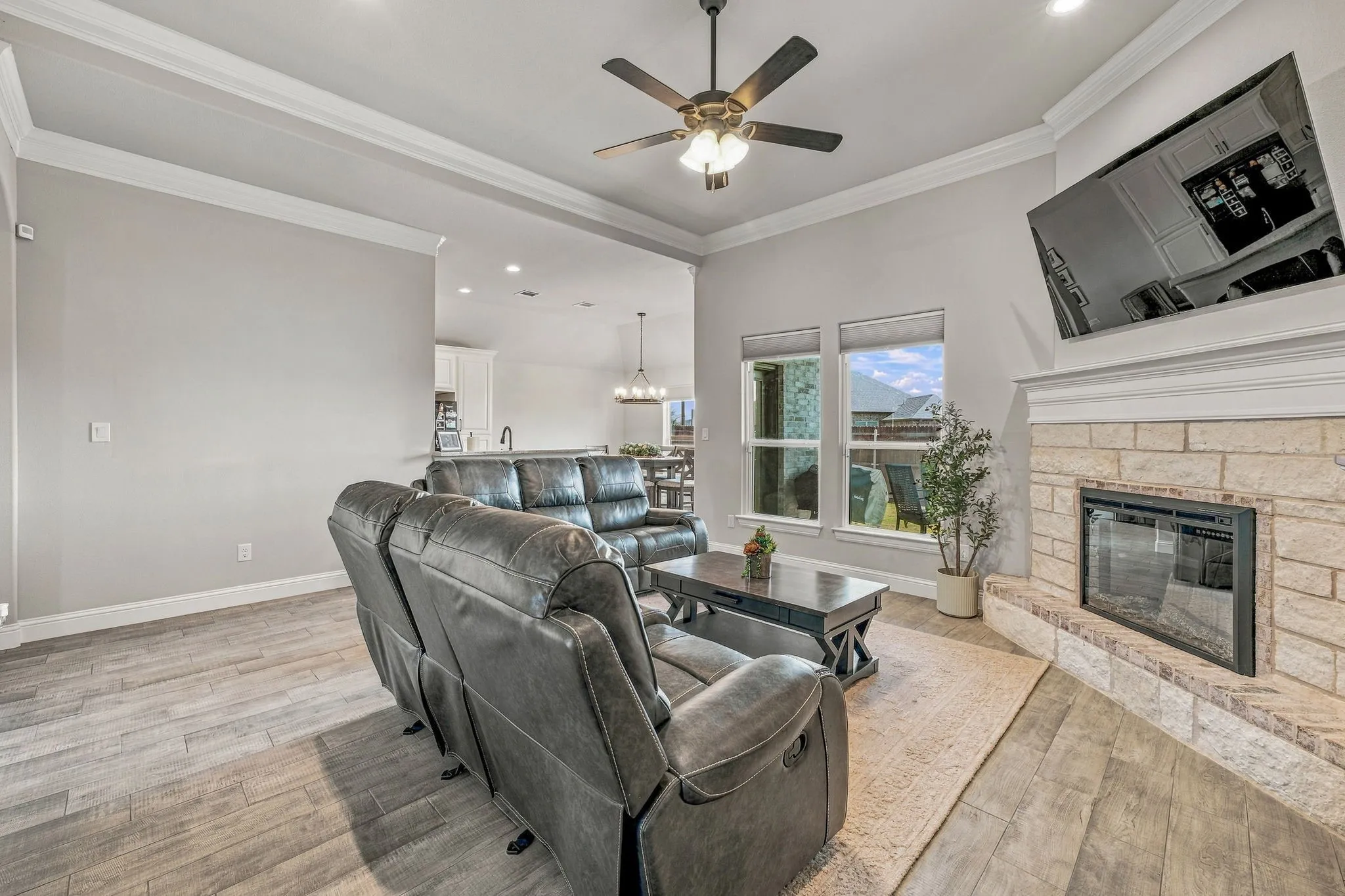 Spacious living area featuring wood-finish flooring, a stone-clad fireplace with a mantel, and crown molding accents