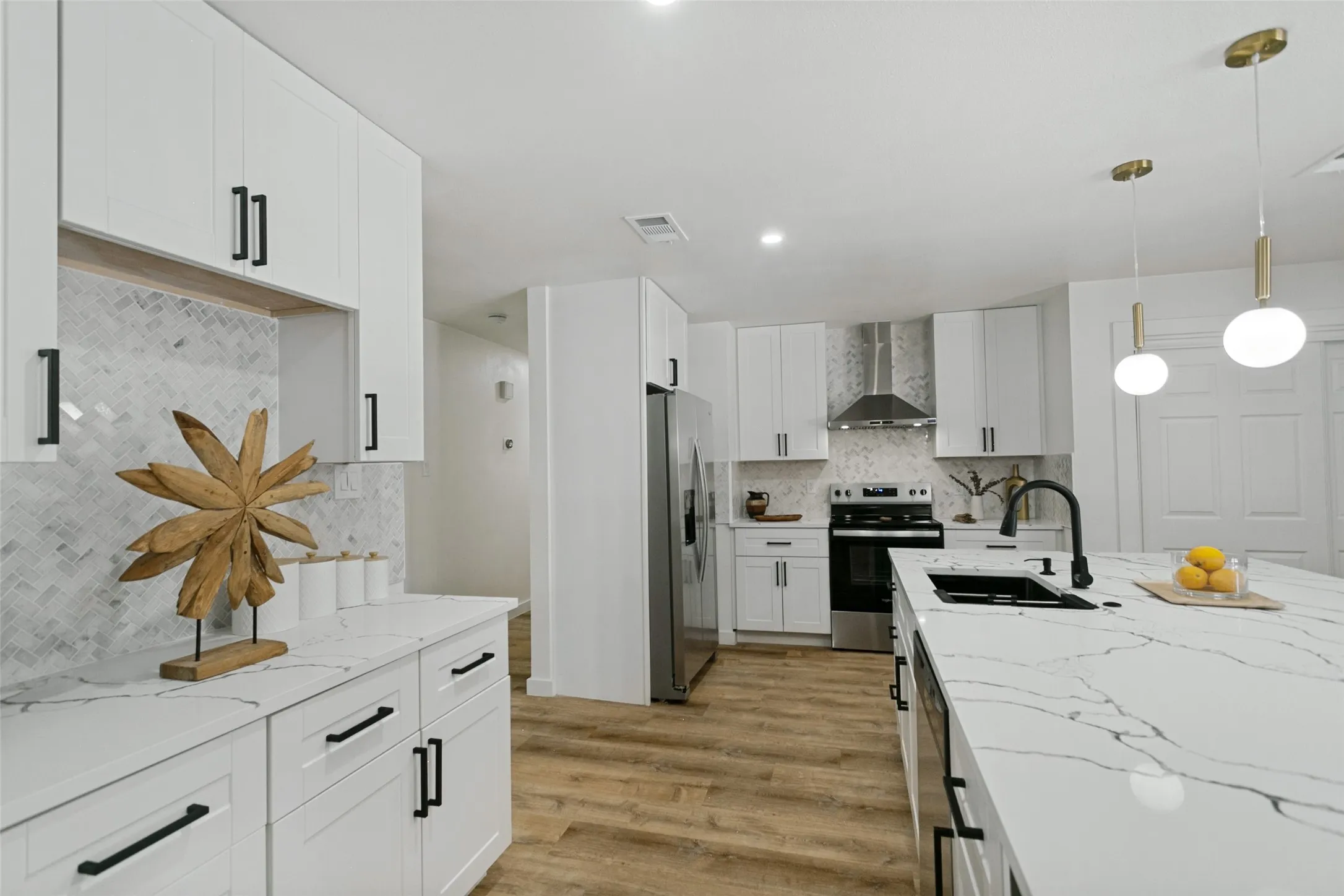 Kitchen featuring white shaker-style cabinetry, a central island with a sink, stainless steel appliances, herringbone-patterned tile backsplash, and wood-finish flooring