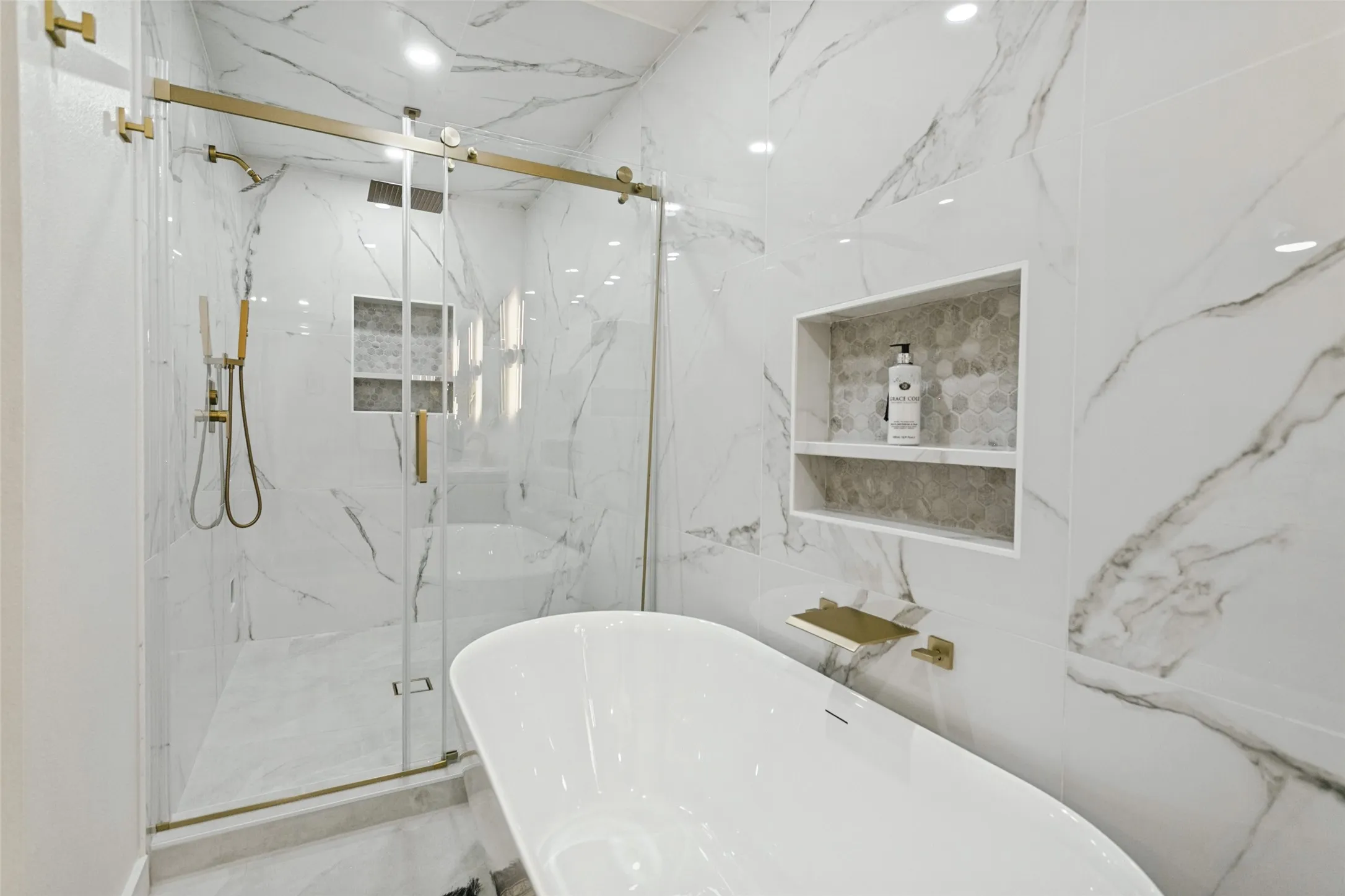 Freestanding soaking tub, large format wall tiling, glass-enclosed shower with gold-tone fixtures, and recessed shelving