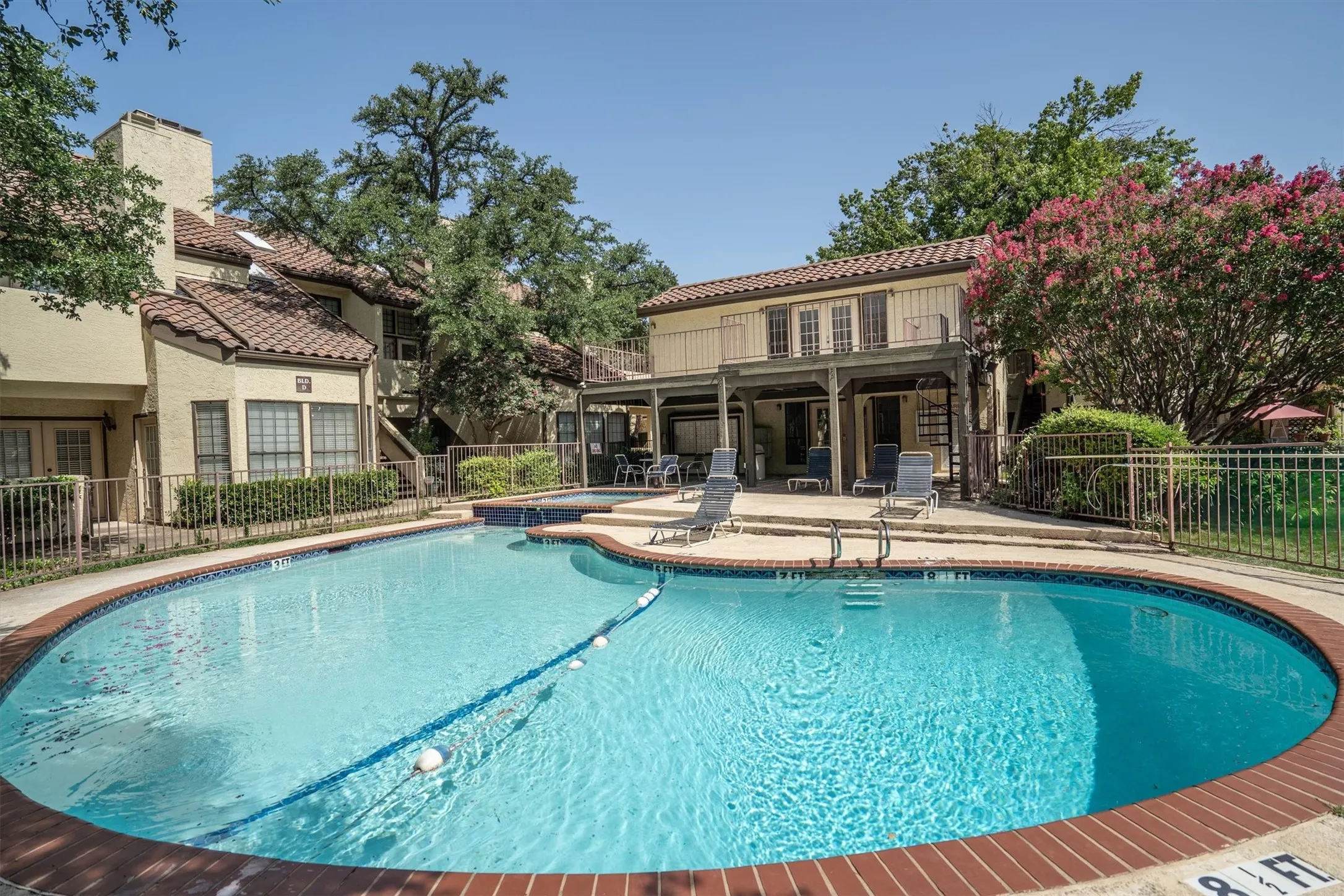 Outdoor swimming pool with brick coping, stucco building exterior, tile roof, and mature trees