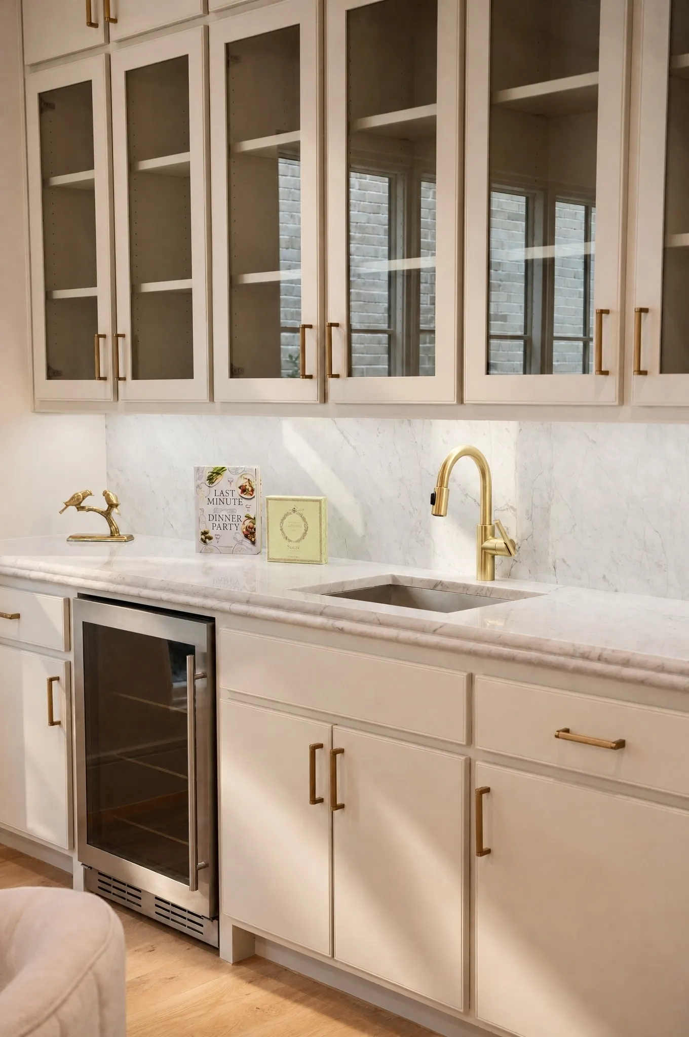 Built-in beverage cooler, undermount sink with gold-tone faucet, white flat-panel cabinetry, light marble countertop, and glass-front upper cabinets