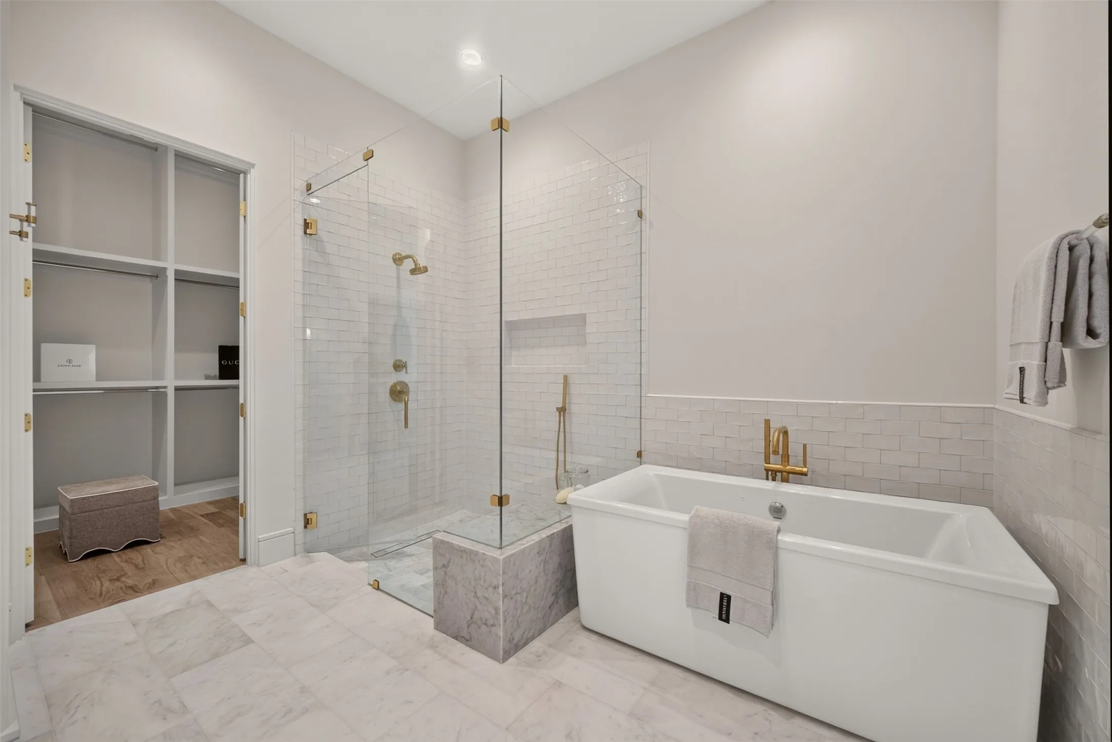 Freestanding soaking tub, glass-enclosed shower with subway tile, built-in shower bench, gold-finish fixtures, and a walk-in closet with built-in shelving