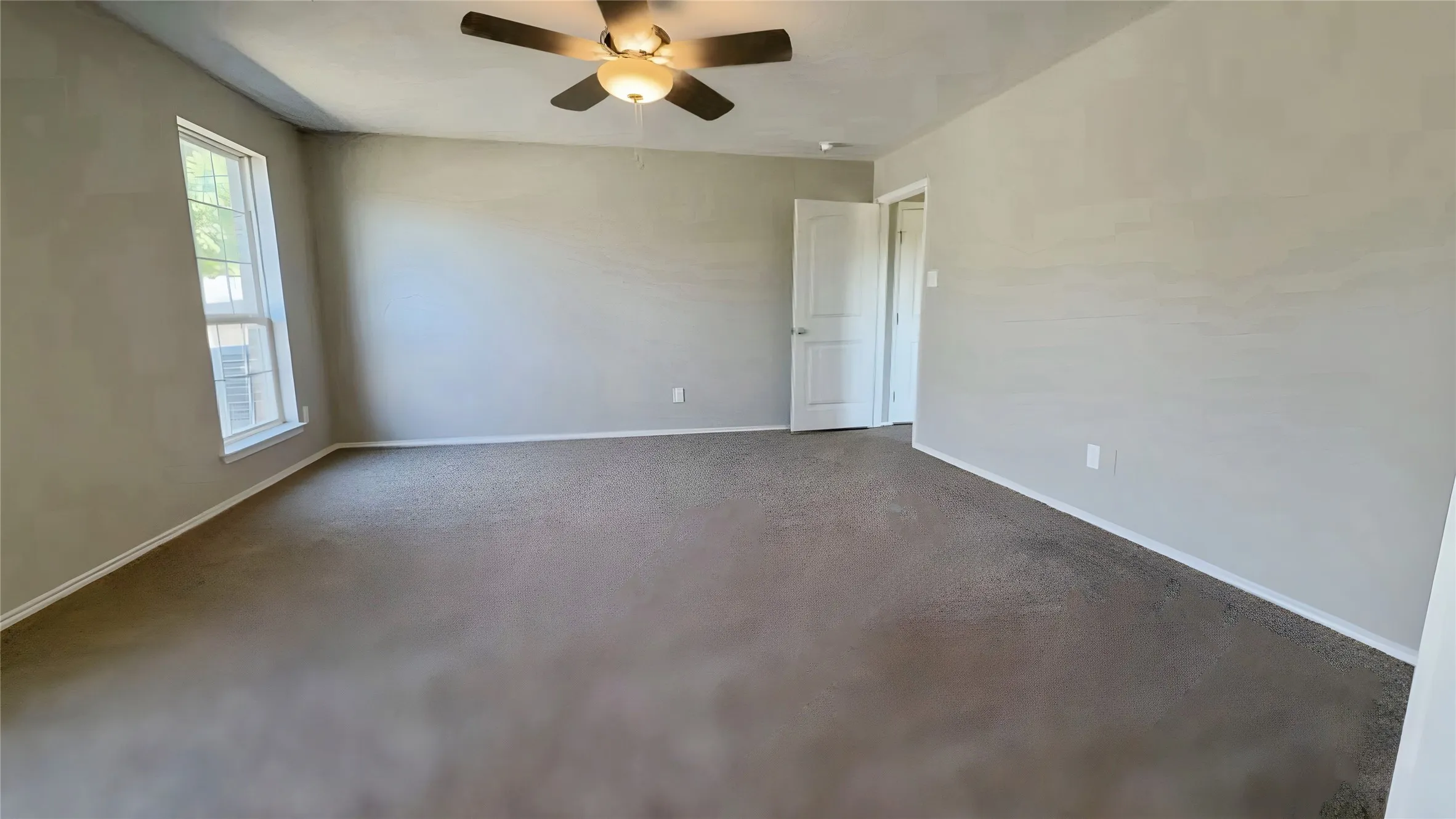 Spacious interior room with carpet flooring, light-toned walls, a ceiling fan, and a window providing natural illumination