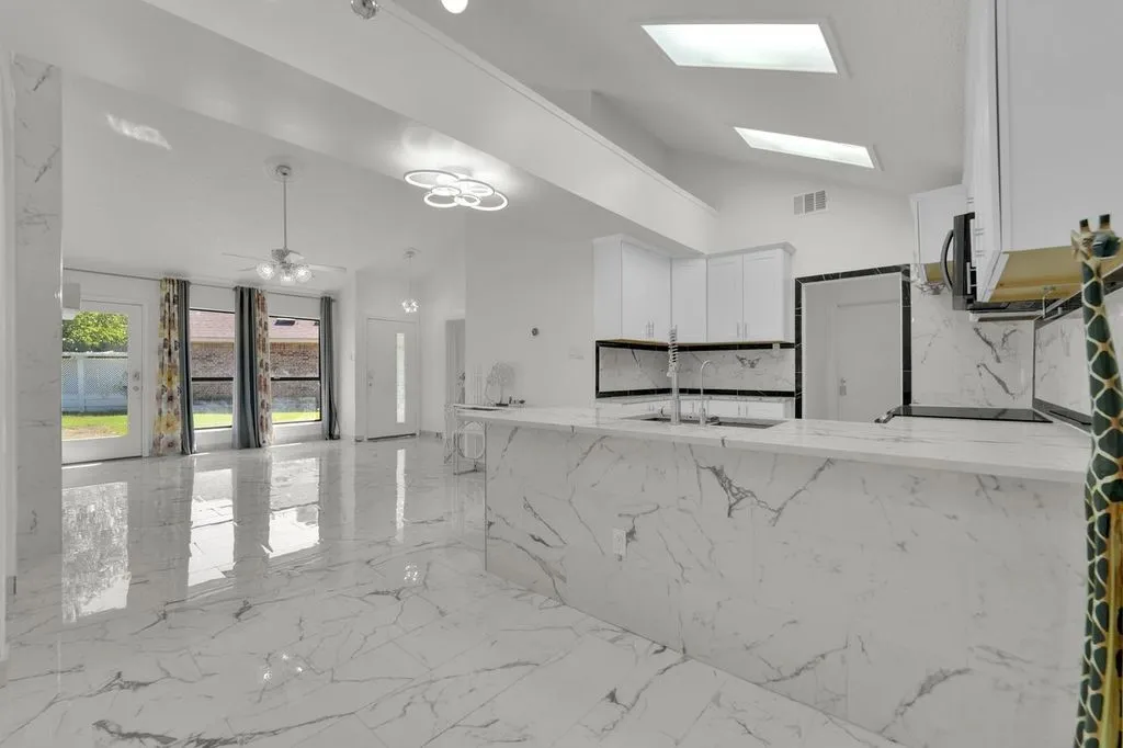 Vaulted ceilings with skylights, white cabinetry, a kitchen island with an integrated sink, and white marble-look flooring