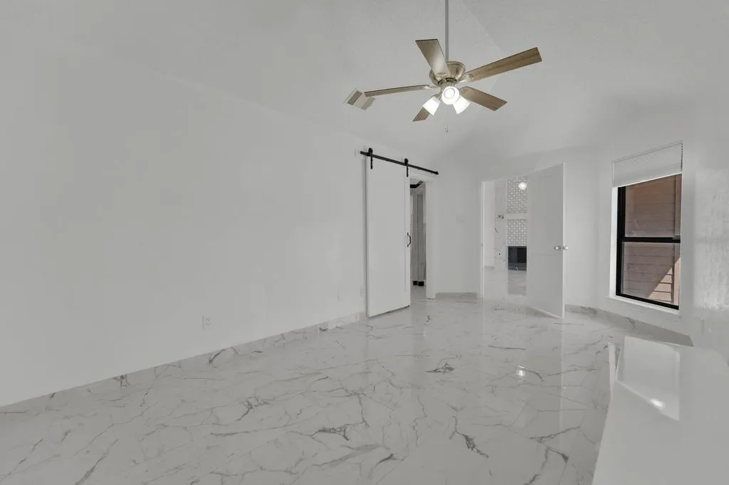Vaulted ceiling with ceiling fan, white walls, marble-look tile flooring, barn door, and a large window