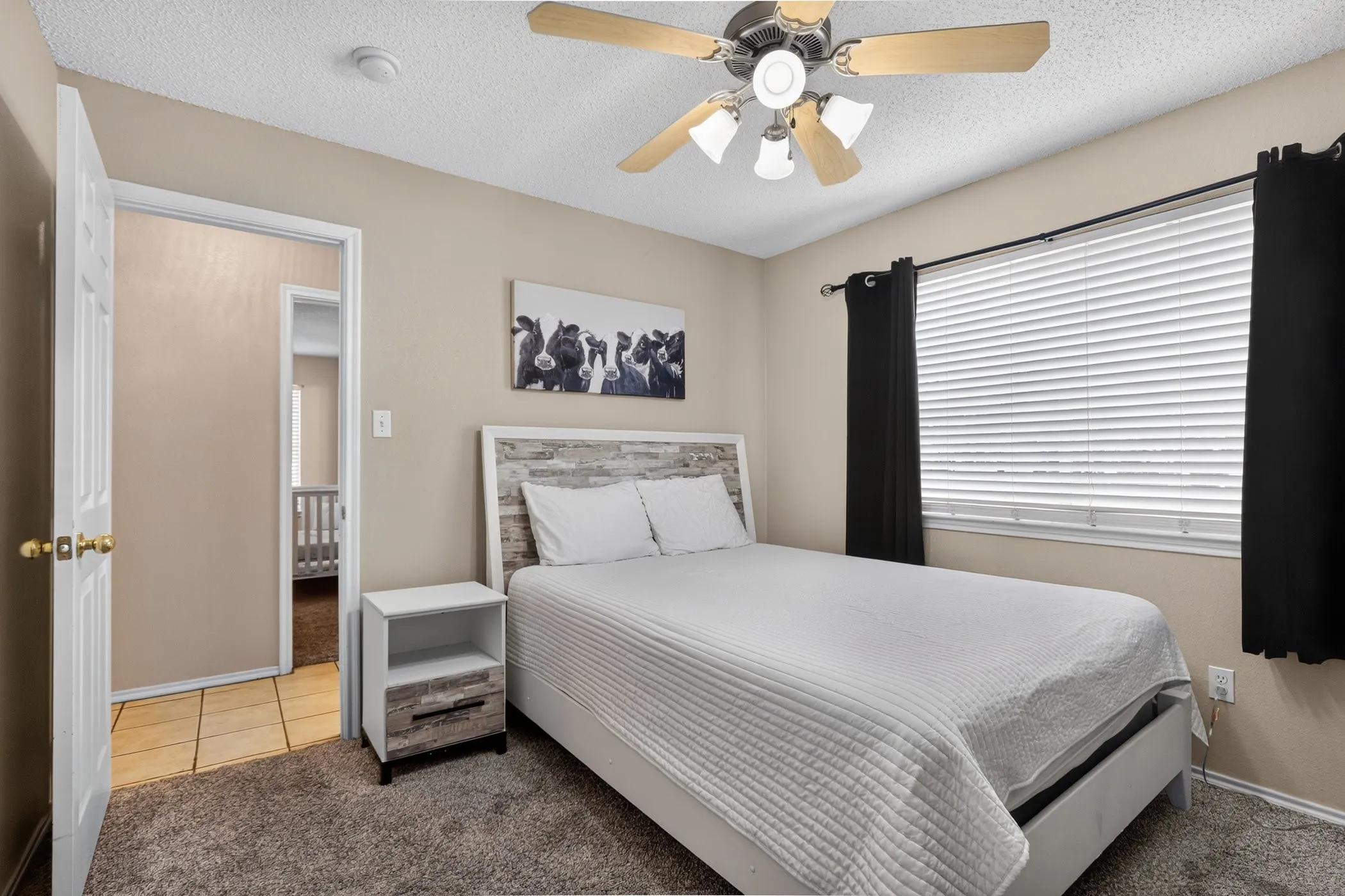 Neutral wall color, ceiling fan with light fixture, window with blinds, carpeted flooring, and a white door with brass hardware