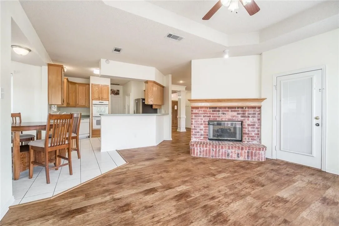 Tray ceiling with a ceiling fan, brick fireplace with a wood mantel, wood-finish flooring, and a kitchen with wood cabinetry
