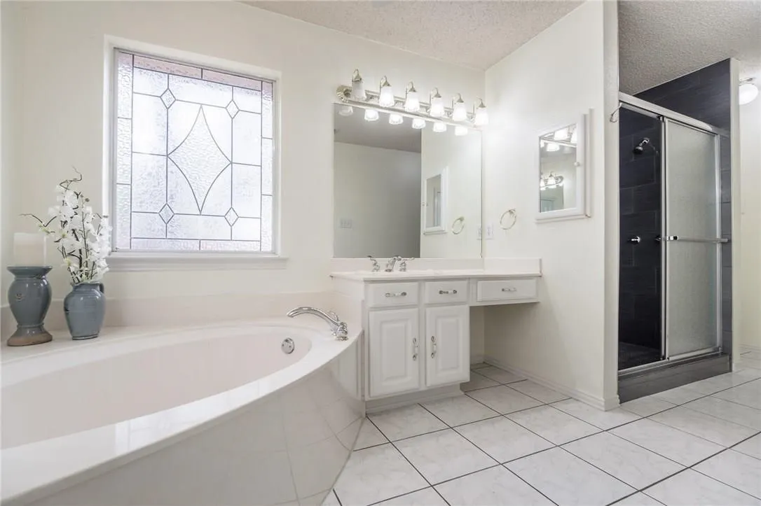White tile flooring, built-in soaking tub, decorative privacy window, dual vanity with white cabinetry, and a glass-enclosed shower
