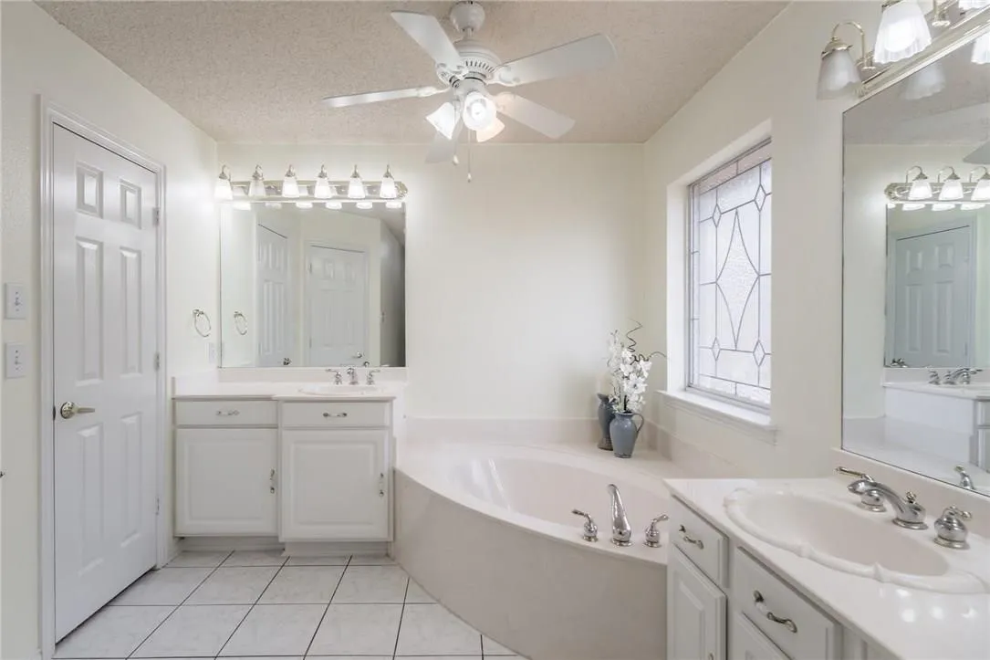Dual vanity sinks, built-in soaking tub, decorative privacy window, ceiling fan with integrated lighting, and tile flooring