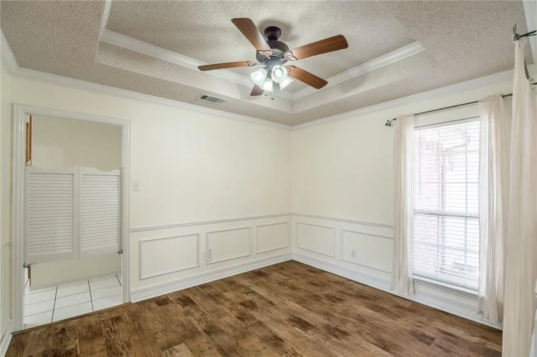 Wood-finish flooring, accent wall paneling, a tray ceiling with a built-in ceiling fan, and a window with blinds