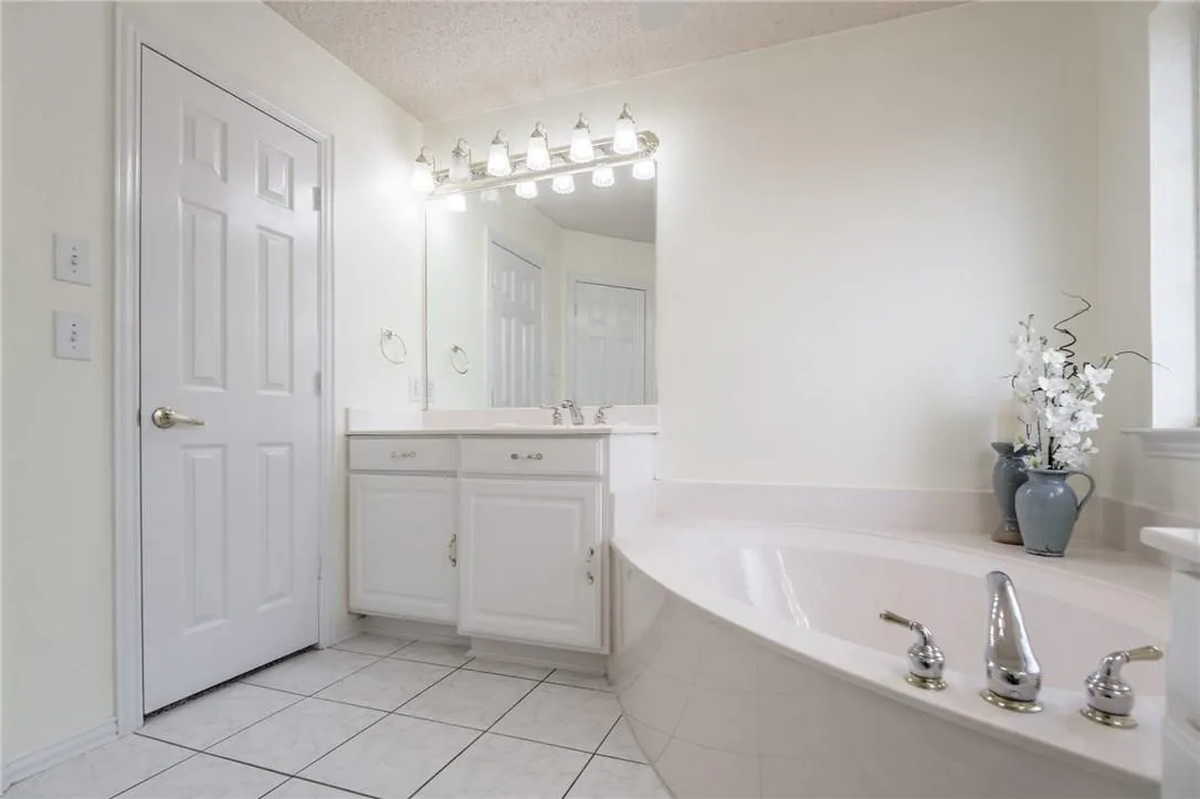 White paneled door, double vanity with white cabinetry, built-in bathtub with deck-mounted chrome fixtures, wall-mounted mirror, and tile flooring