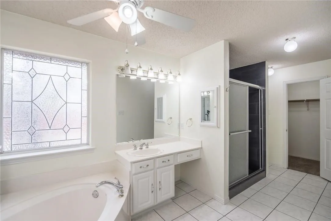 Bathroom featuring a built-in tub, white vanity with integrated sink, glass-enclosed shower, decorative privacy window, and ceiling fan