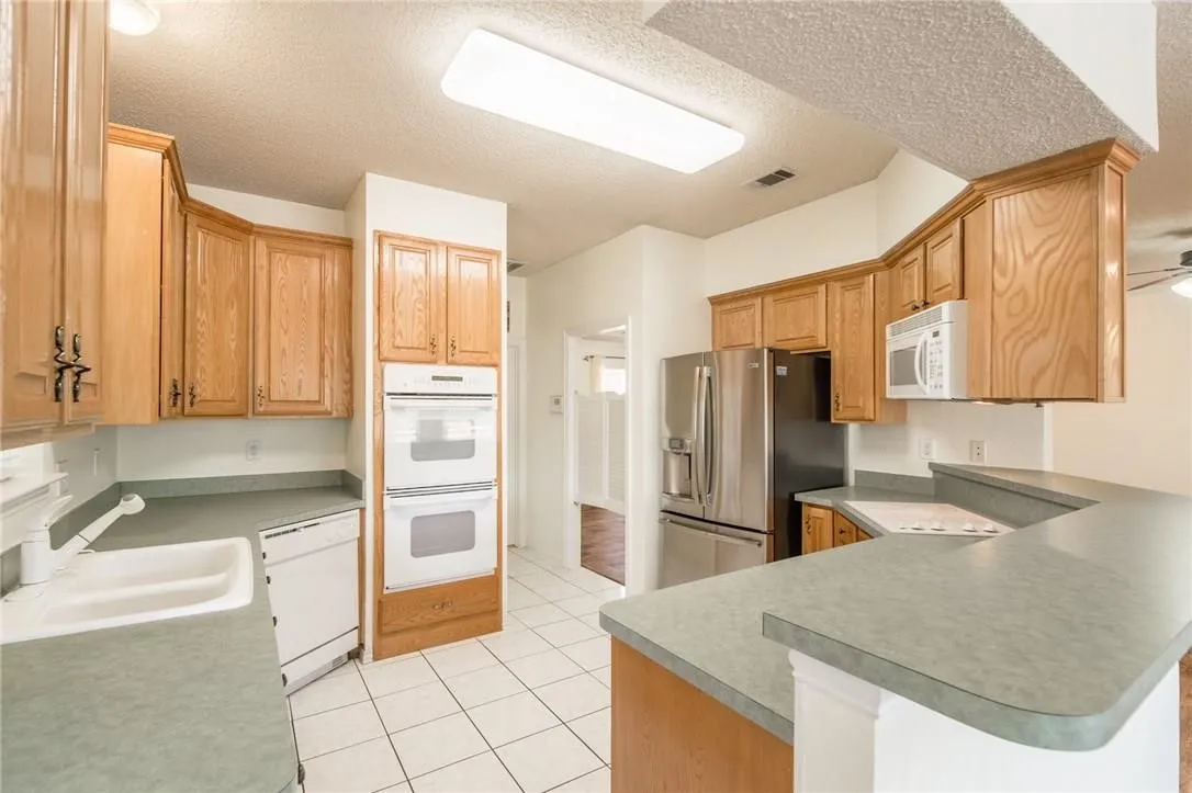 Kitchen featuring wood cabinetry, white tile flooring, stainless steel refrigerator, white double wall ovens, and a built-in cooktop