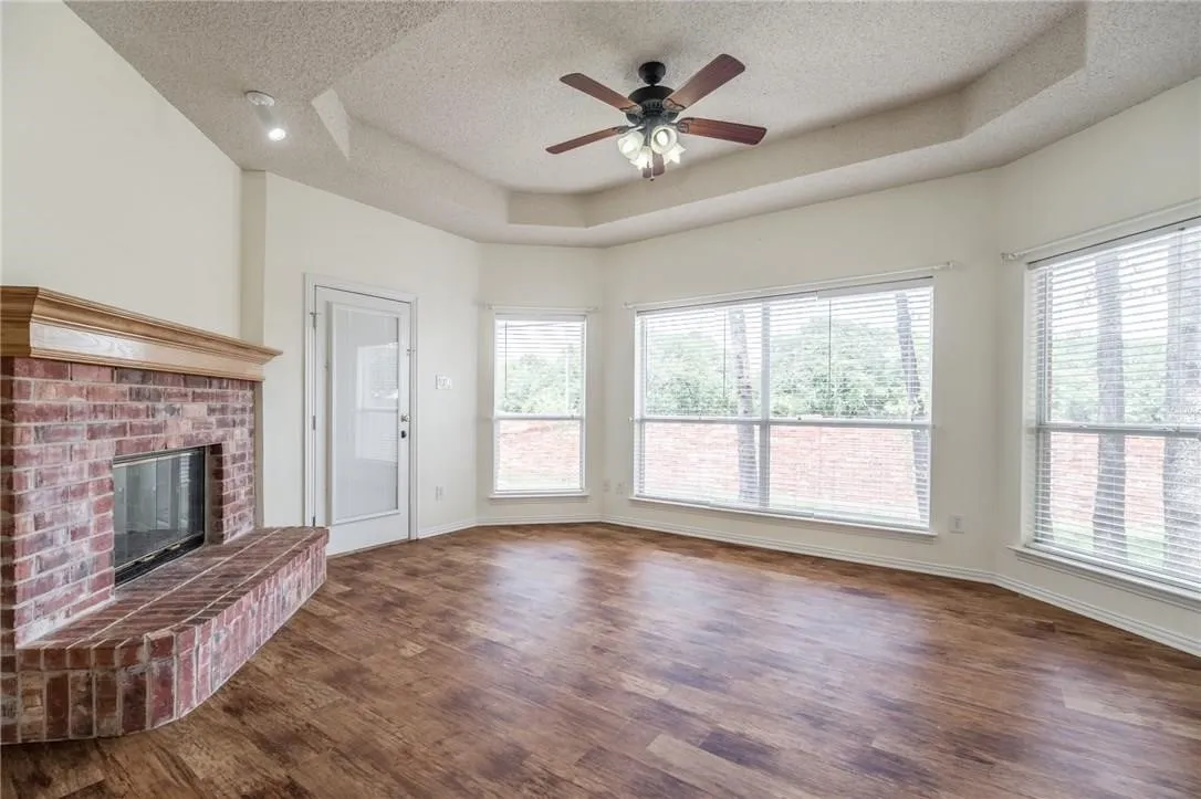 Wood-finish flooring throughout, brick fireplace with wood mantel, multiple windows with blinds, tray ceiling, and ceiling fan with integrated lighting