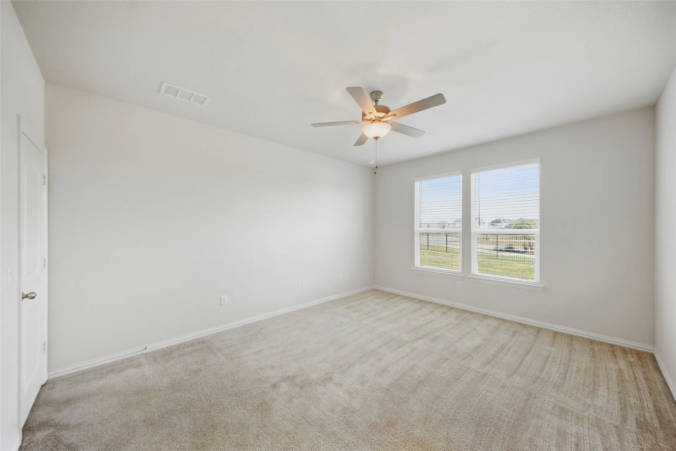 Spacious room featuring neutral carpeting, two large windows with blinds, a ceiling fan with integrated lighting, and a white paneled door