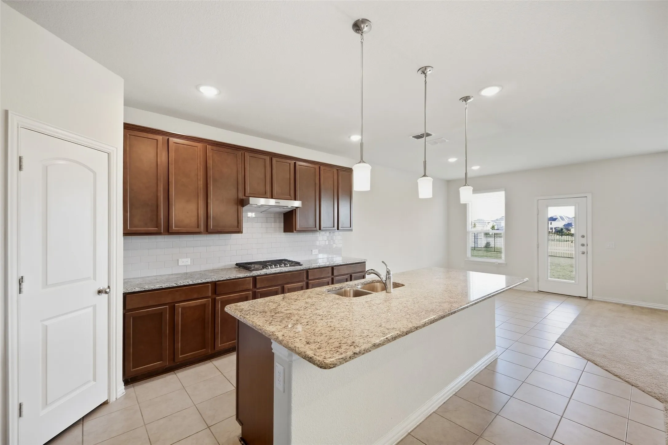 Kitchen featuring wood cabinetry, a granite-finish island with a double basin sink, stainless steel gas cooktop, tile backsplash, and pendant lighting