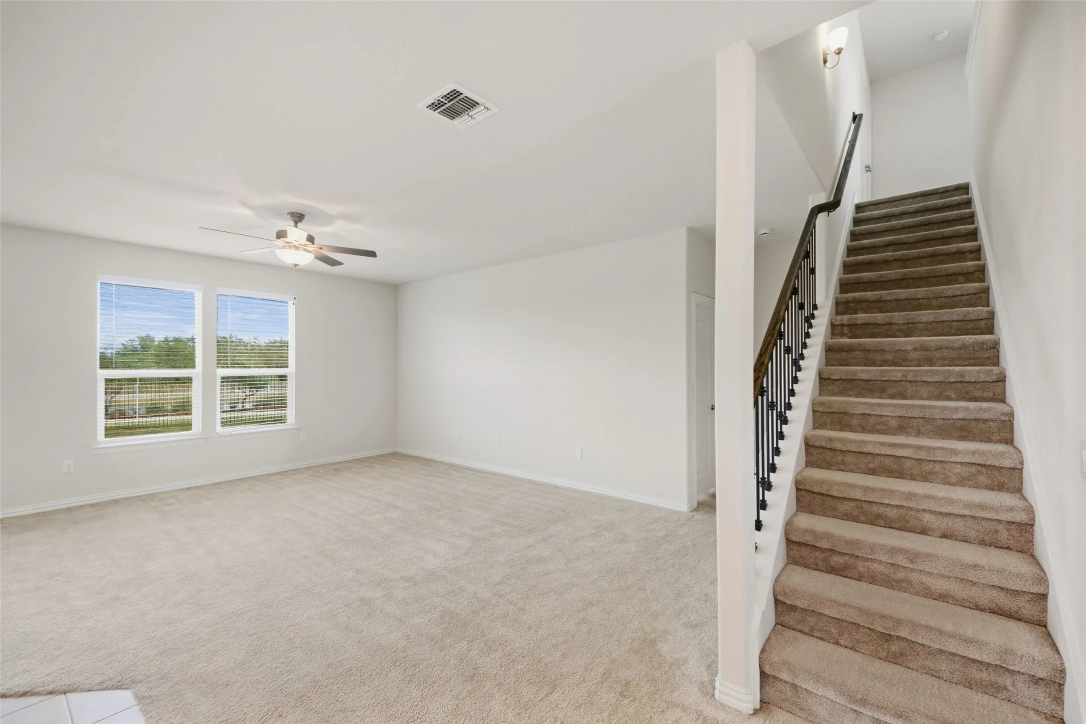 Open concept living area featuring dual windows, a ceiling fan with integrated lighting, and a carpeted staircase with dark iron balusters