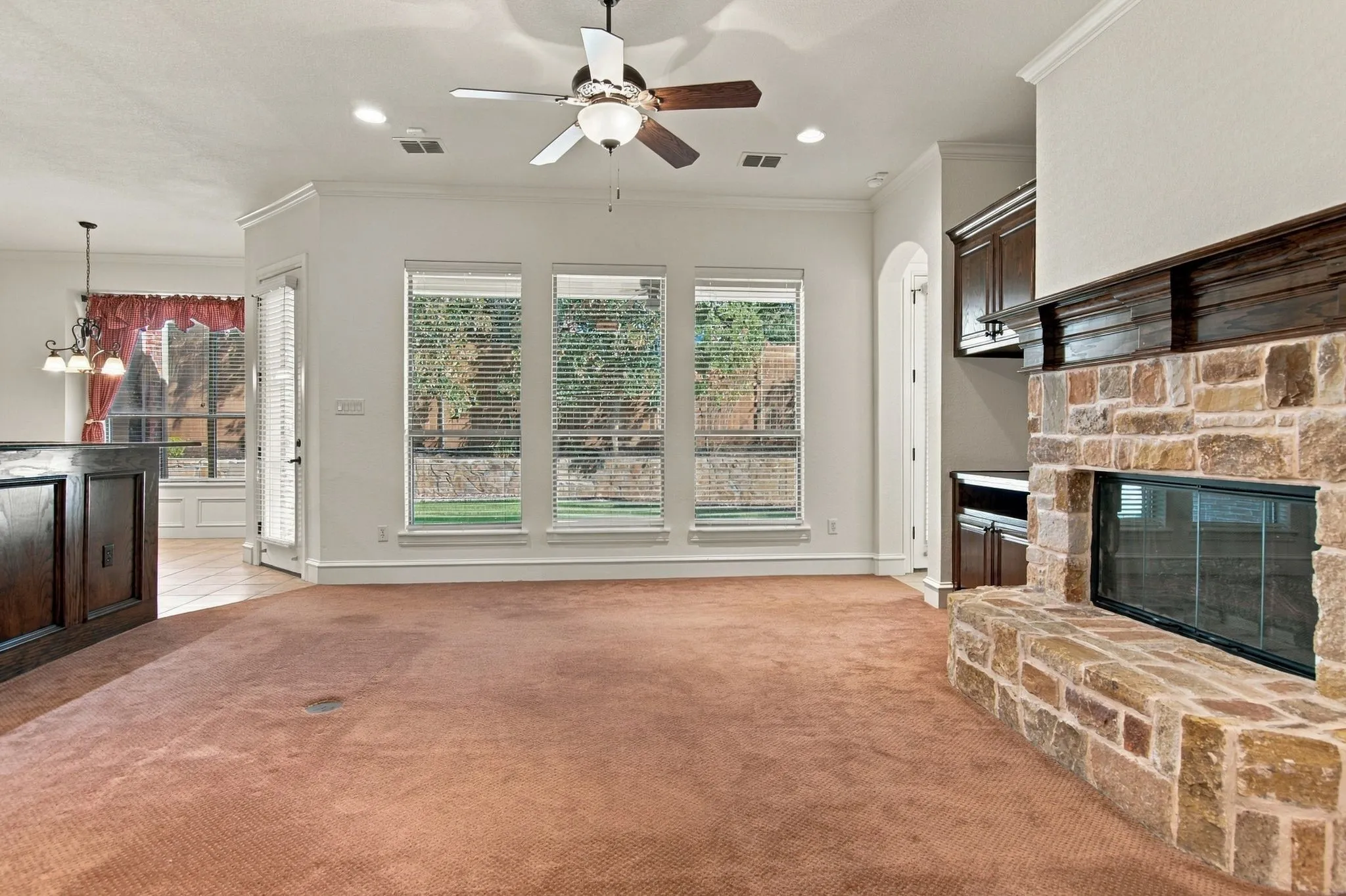 Stone-clad fireplace with dark wood mantel, ceiling fan with integrated lighting, recessed lighting, multiple windows with horizontal blinds, and built-in wood cabinetry