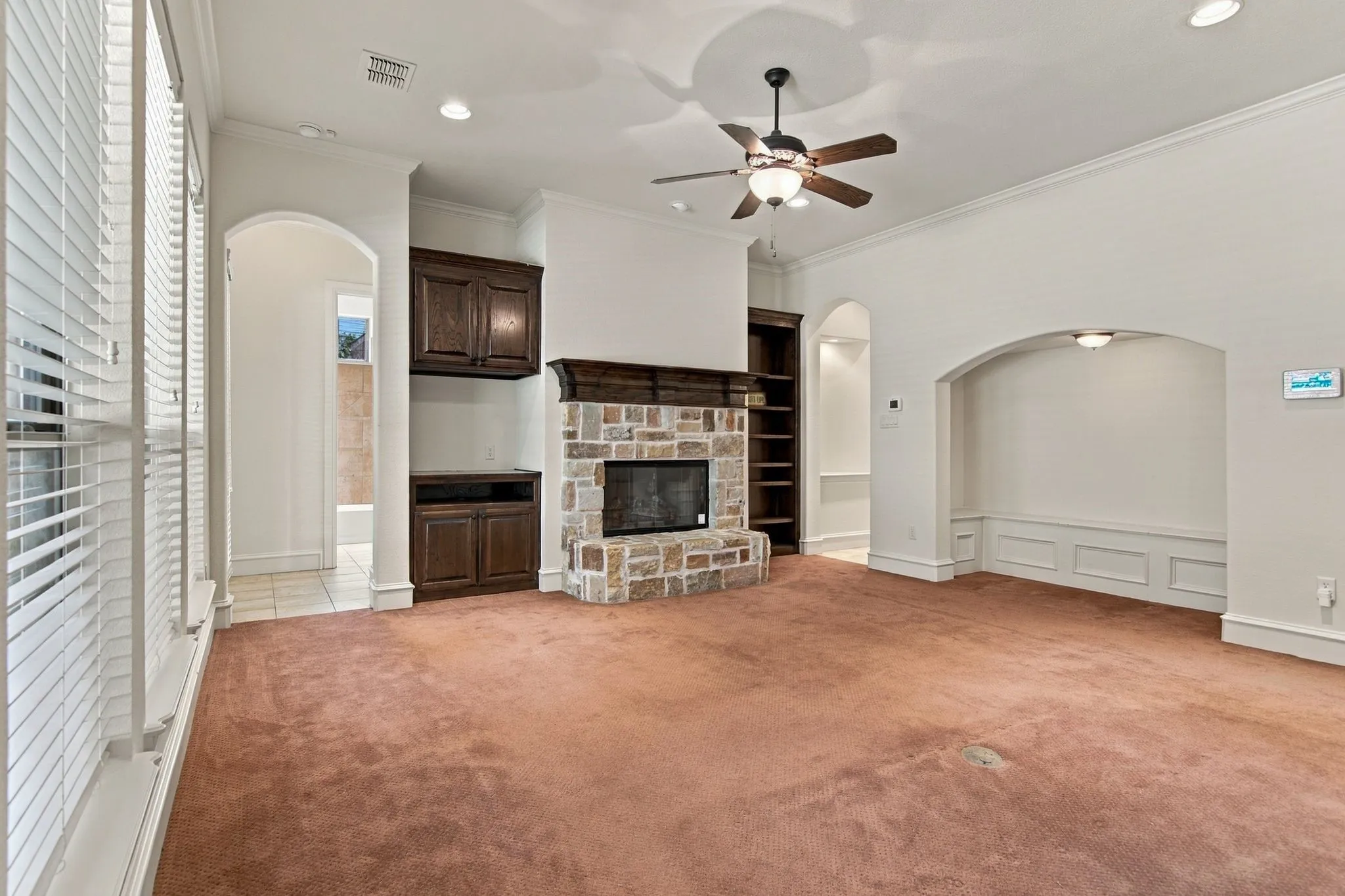 Arched entryways, built-in shelving, stone-faced fireplace with wood mantel, built-in dark wood cabinetry, and a ceiling fan with integrated lighting