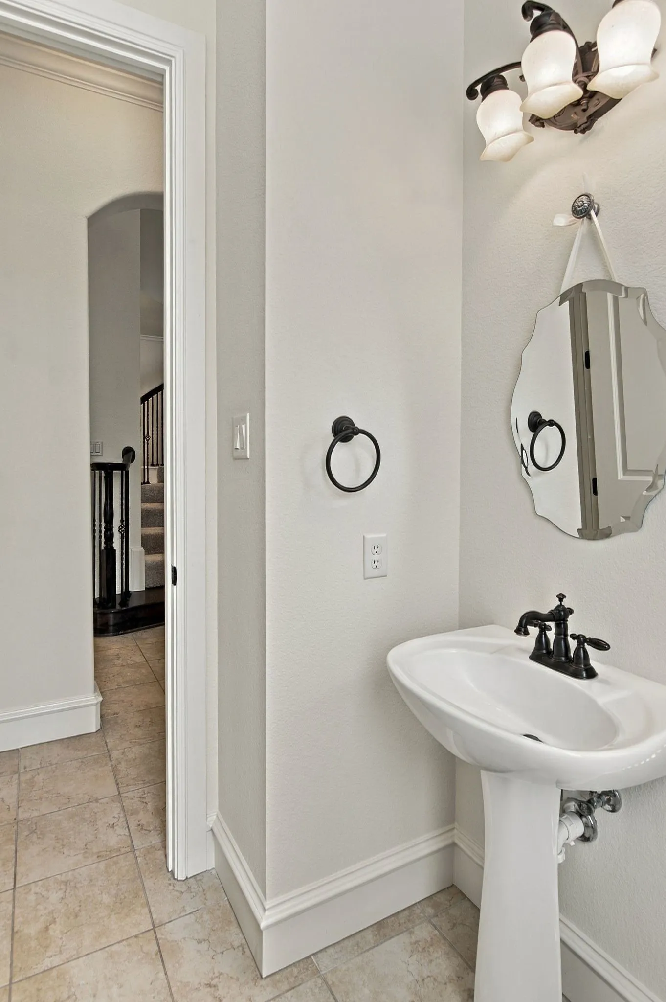 Pedestal sink with black-finish faucet, decorative wall mirror, three-light vanity fixture, and tiled flooring