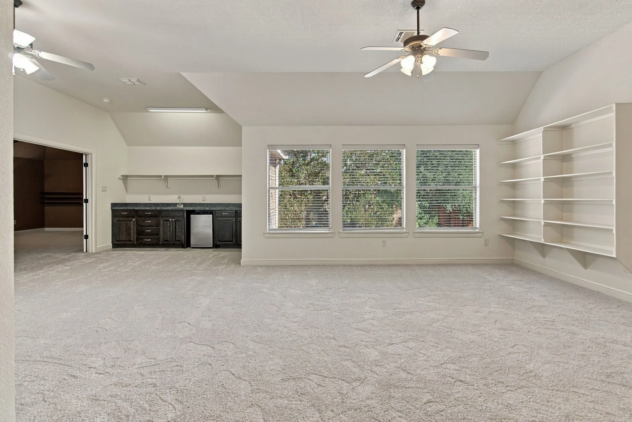 Expansive room featuring light-colored carpeting, multiple windows, built-in shelving, and a wet bar with dark cabinetry and a compact refrigerator