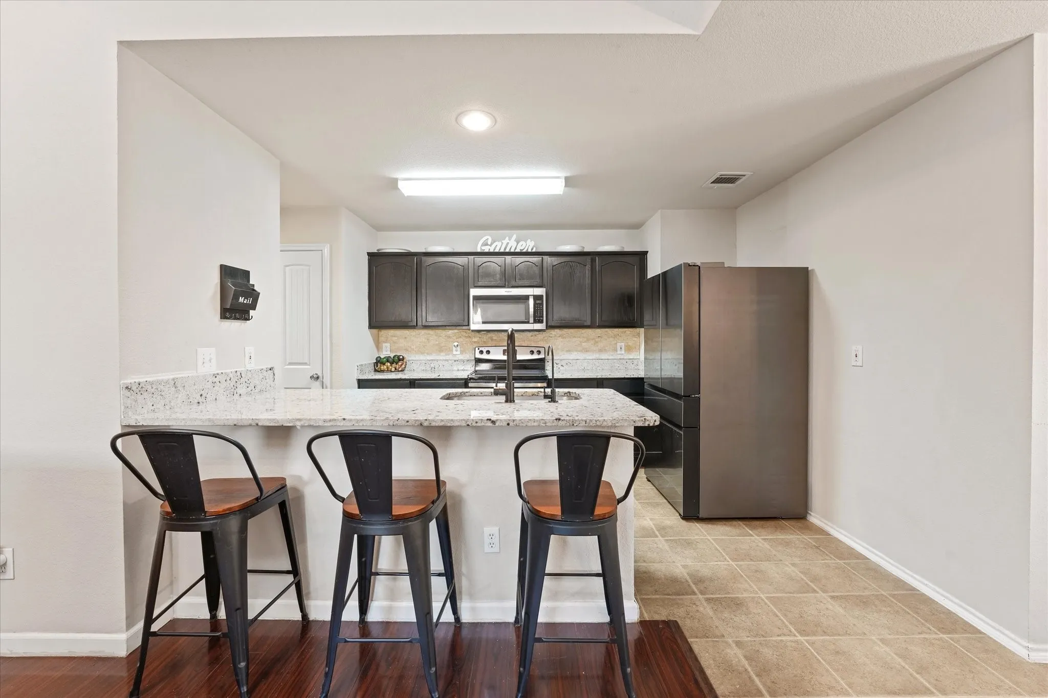 Kitchen featuring a peninsula with a light-colored countertop, dark wood-finish cabinetry, stainless steel microwave, black refrigerator, and tile flooring