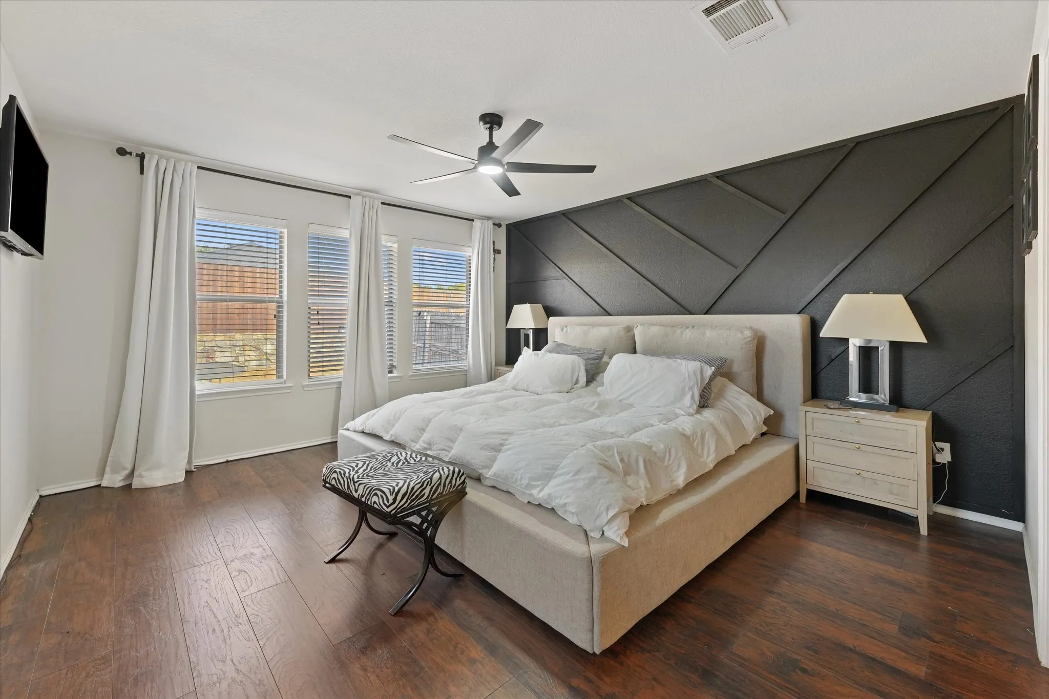 Wood-finish flooring, accent wall with geometric paneling, multiple windows with blinds, ceiling fan with integrated lighting