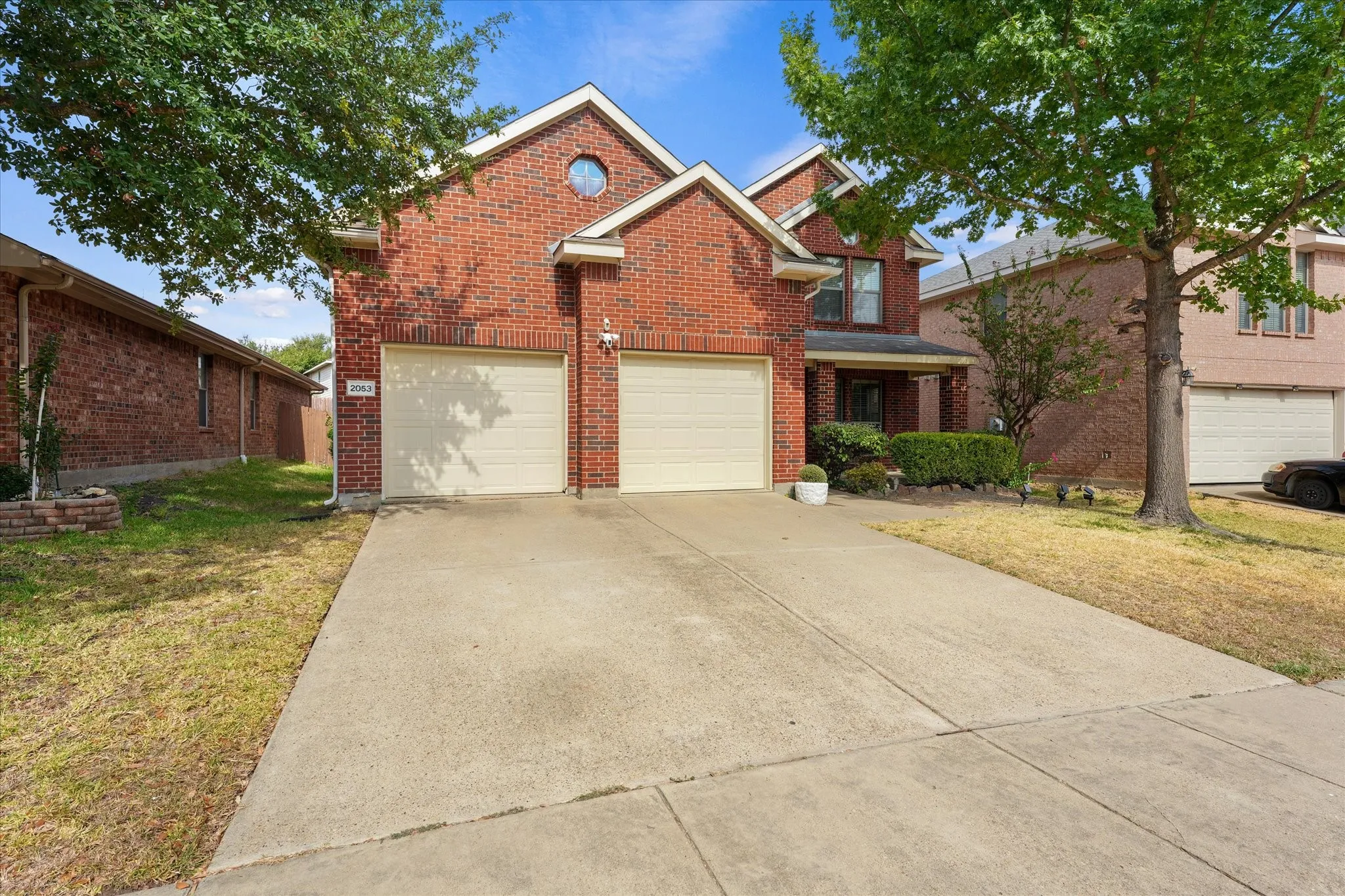 Brick facade residence featuring a two-car garage, concrete driveway, front porch, and mature trees