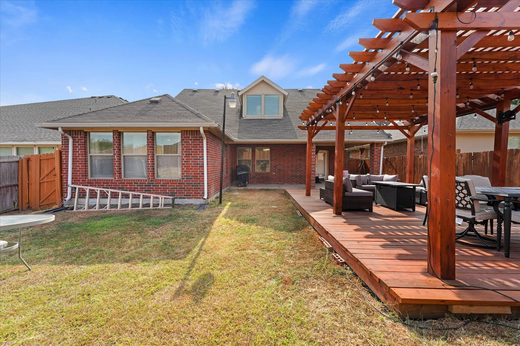 Expansive wood-finish deck with an attached pergola featuring string lighting, complemented by a brick exterior with multiple windows