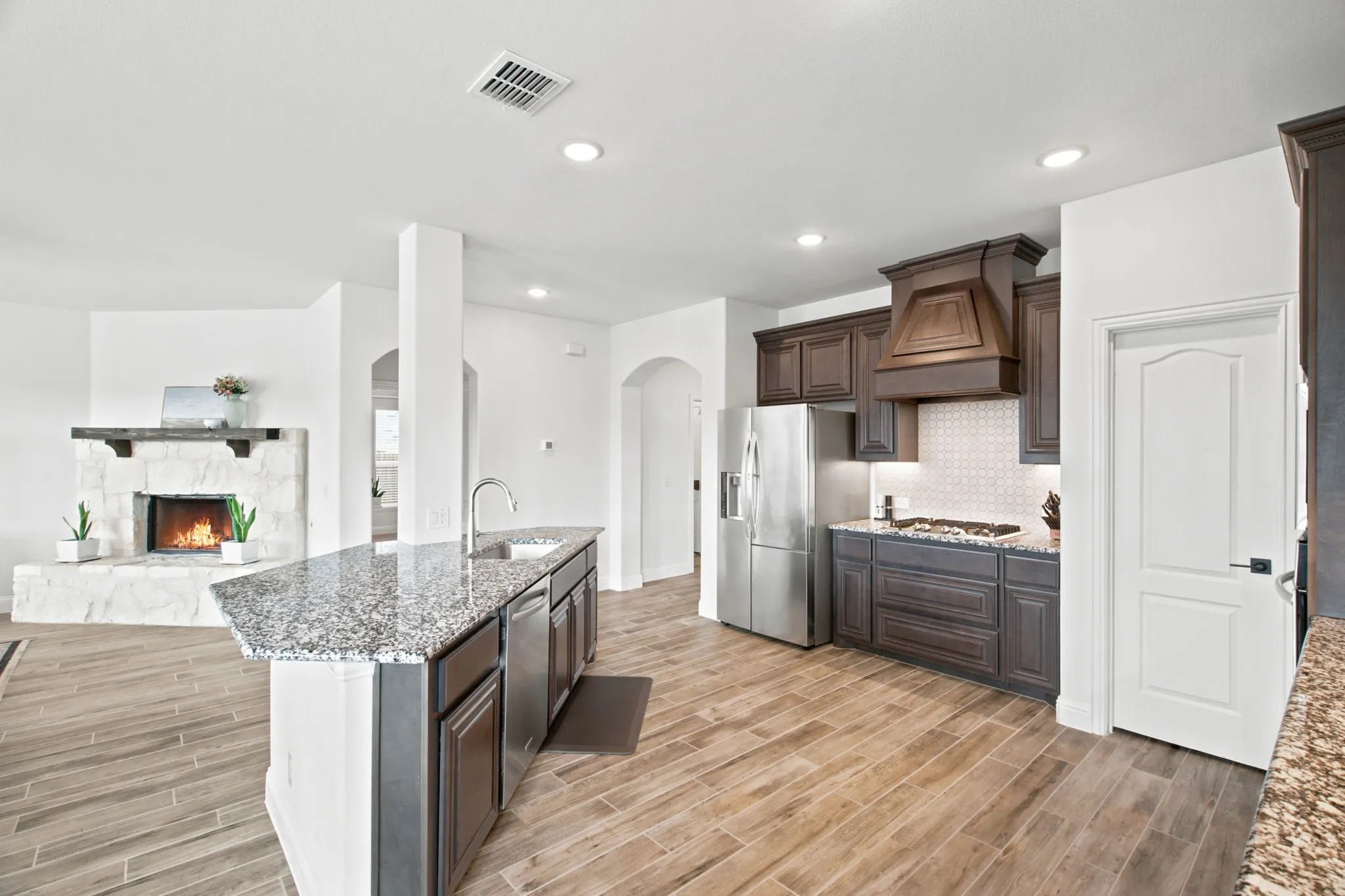 Open-concept design featuring wood-look tile flooring, a kitchen island with stone countertops, stainless steel appliances, and a stone-faced fireplace