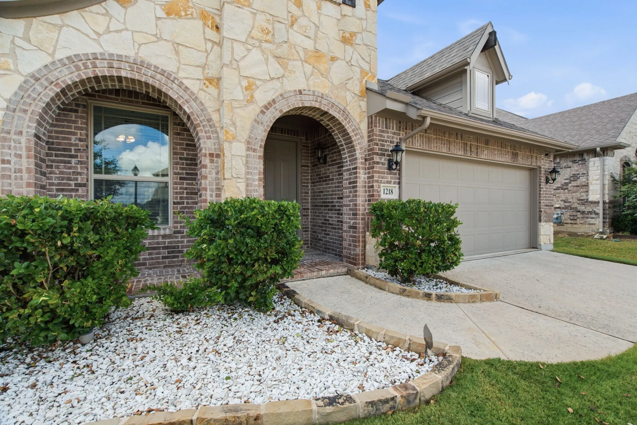 Stone and brick facade with arched entryway, concrete driveway, attached garage, and landscaping with white stone accents