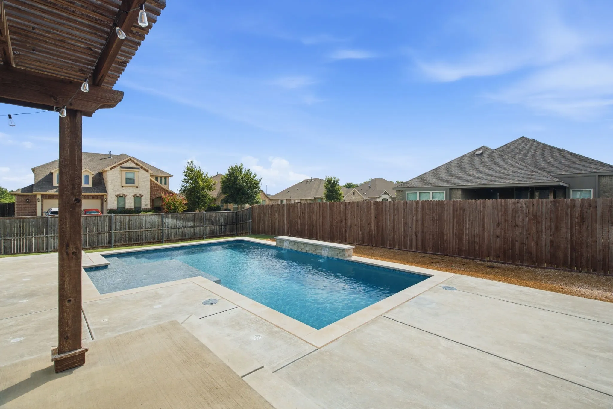 Outdoor swimming pool with freestanding spa feature, surrounding concrete decking, wood pergola, and privacy fencing