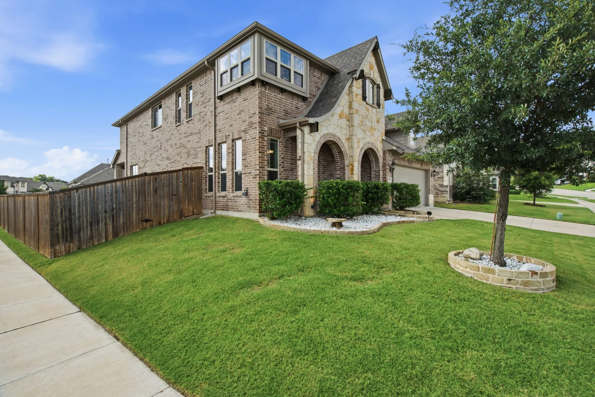 Brick and stone facade, arched entryway, bay window, landscaped front yard with decorative stone beds, and a paved sidewalk