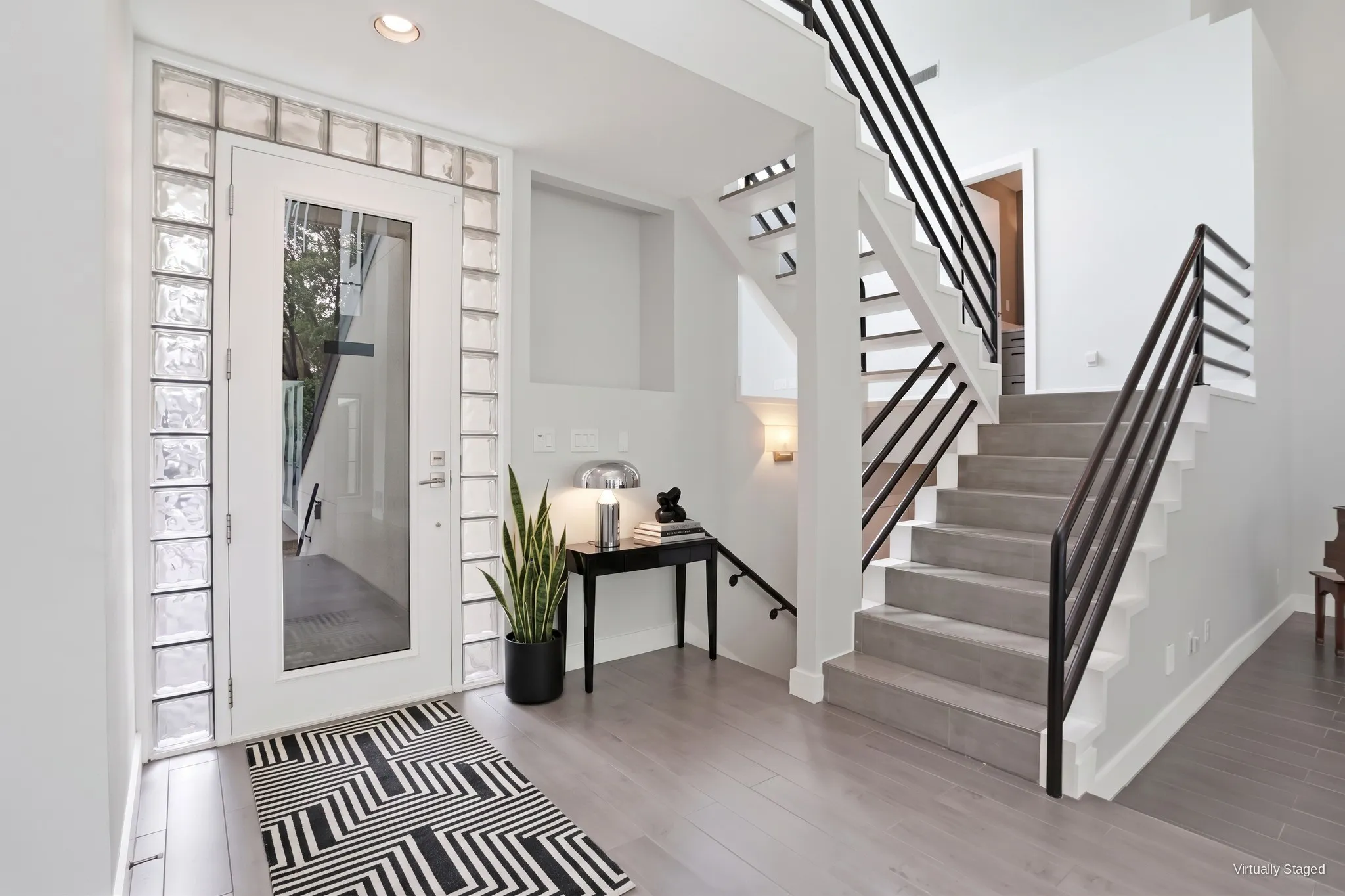 Entry with glass-block sidelights and transom, a full-glass front door, and a floating staircase with black metal railings. Virtually staged.