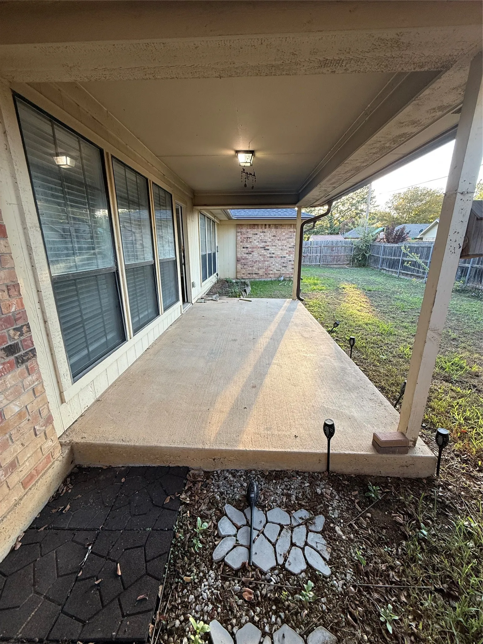 Covered concrete patio with overhead lighting, brick exterior, and a fenced backyard