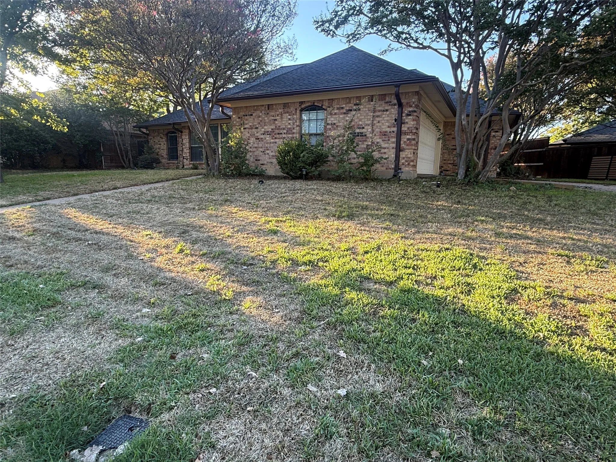 Brick exterior, dark shingle roof, attached garage, established trees, and front yard