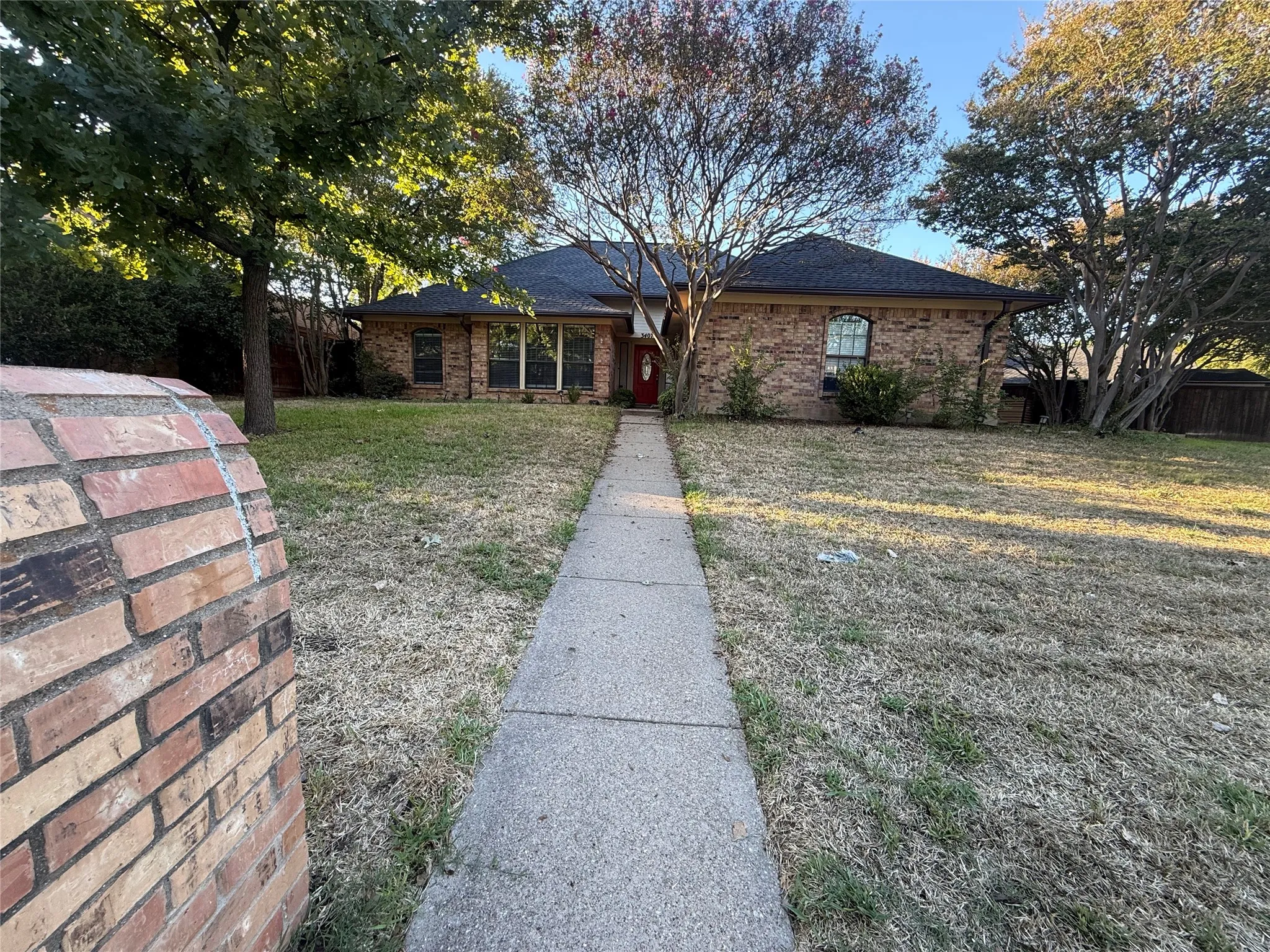 Brick exterior with a red entry door, framed by mature trees, a concrete pathway, and an expansive front lawn