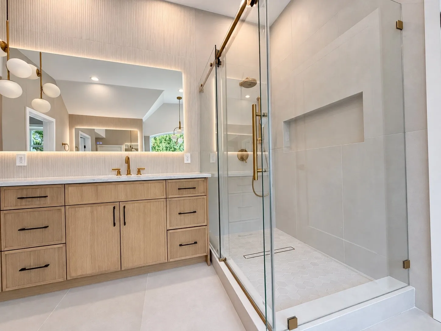 Bathroom featuring a wood-finish vanity with a light-colored countertop, an illuminated mirror, a glass-enclosed shower with gold-tone fixtures, and a tiled shower niche