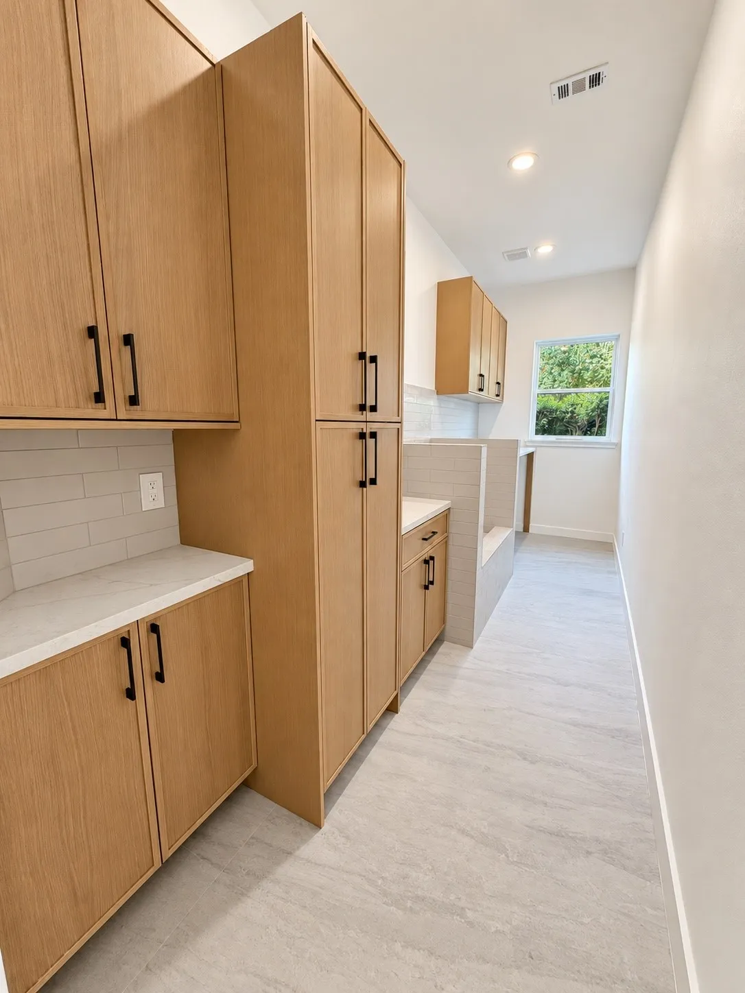 Light wood-finish cabinetry with black hardware, white countertops, tile backsplash, and light gray tile flooring
