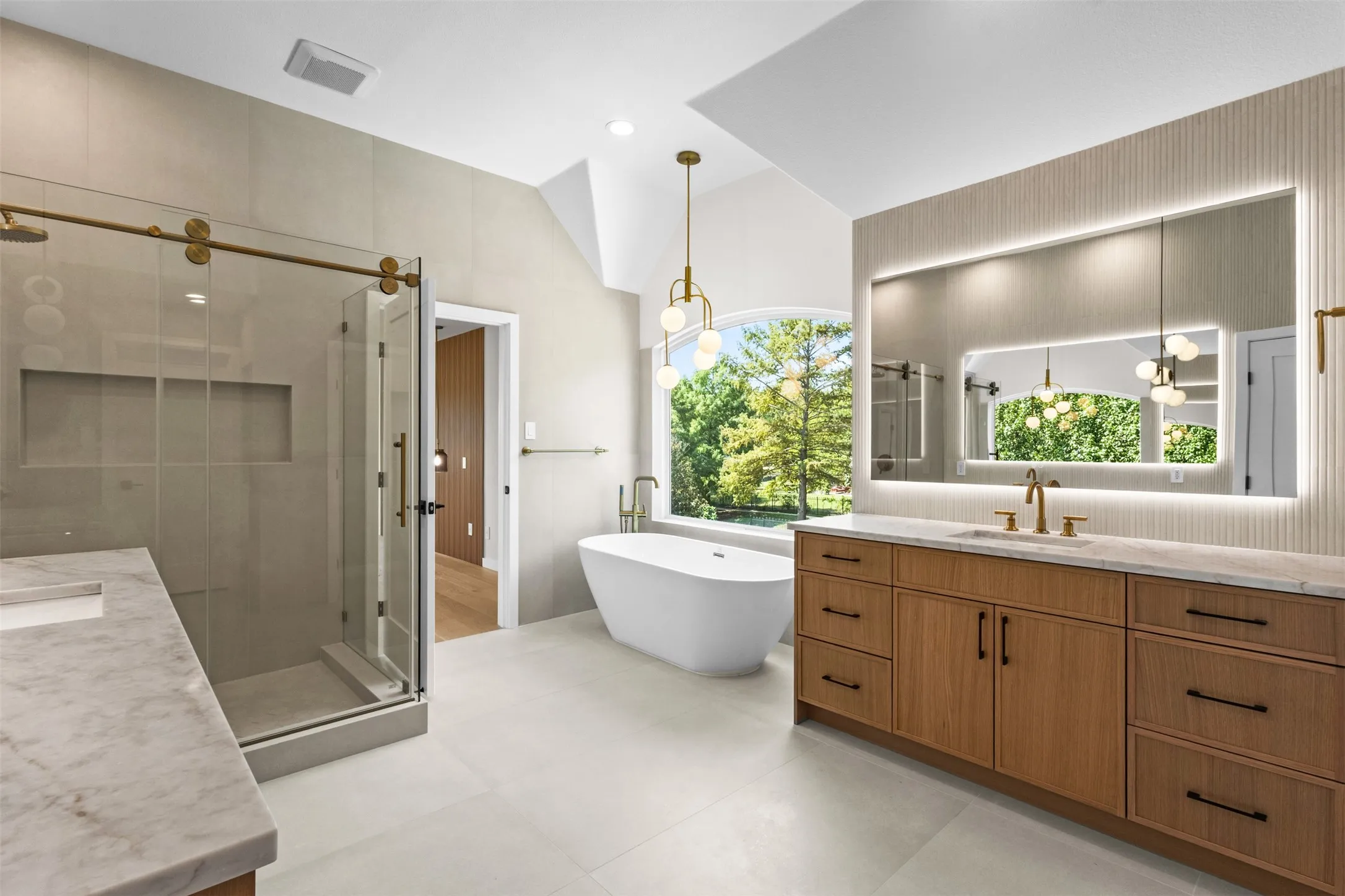 Freestanding soaking tub, dual vanity with wood-finish cabinetry, illuminated medicine cabinet, walk-in shower with glass enclosure, and vaulted ceiling