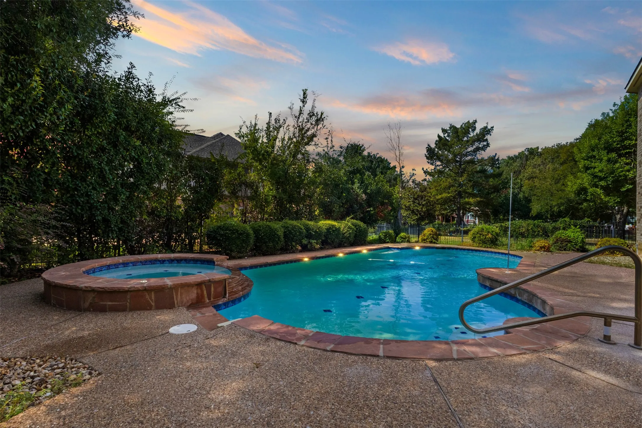 Curved swimming pool with an integrated spa, expansive aggregate concrete decking, and lush perimeter landscaping