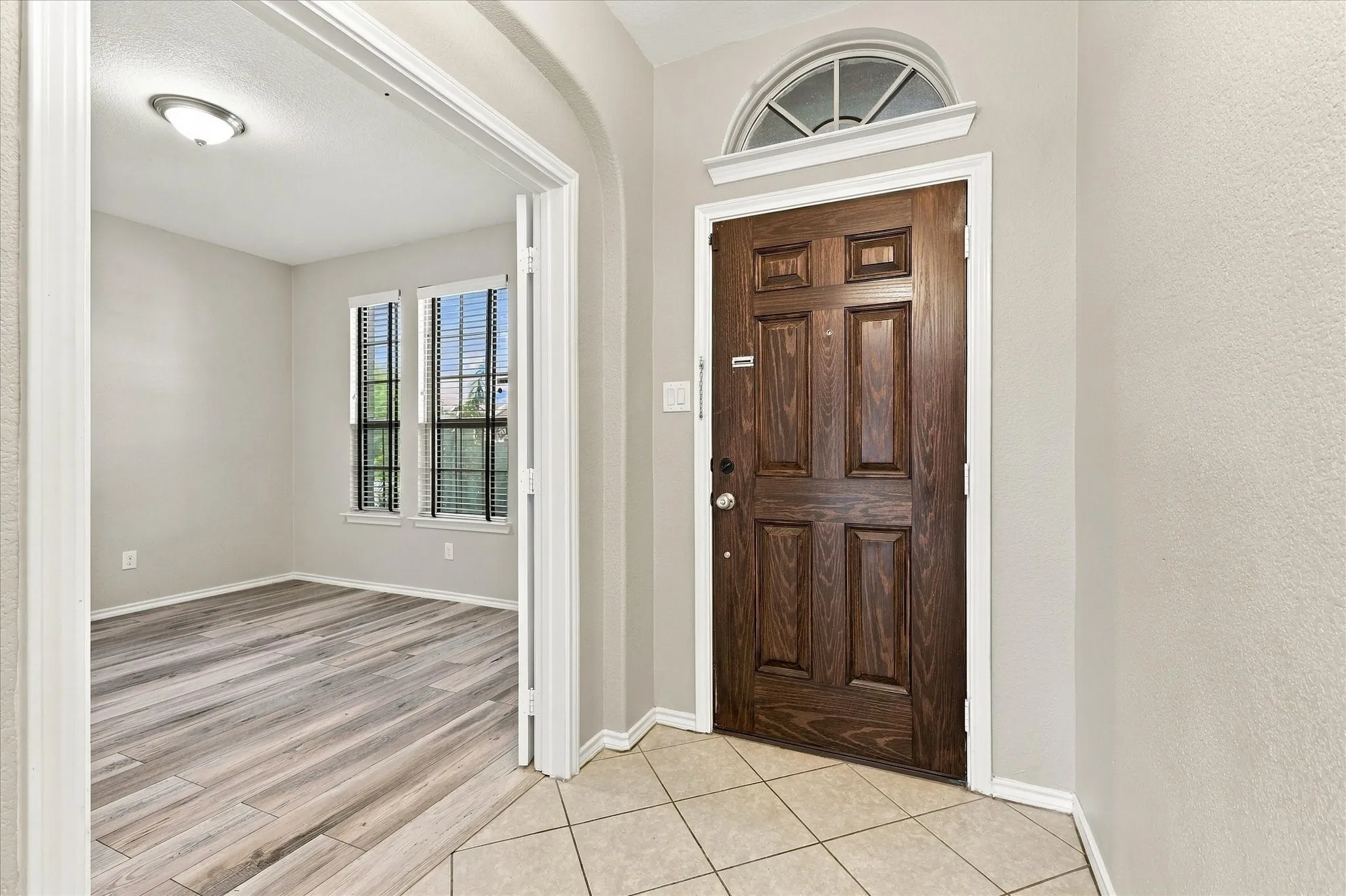 Wood-paneled entry door with an arched transom window, ceramic tile flooring, and a recessed doorway leading to a room with wood-finish flooring and two windows with blinds